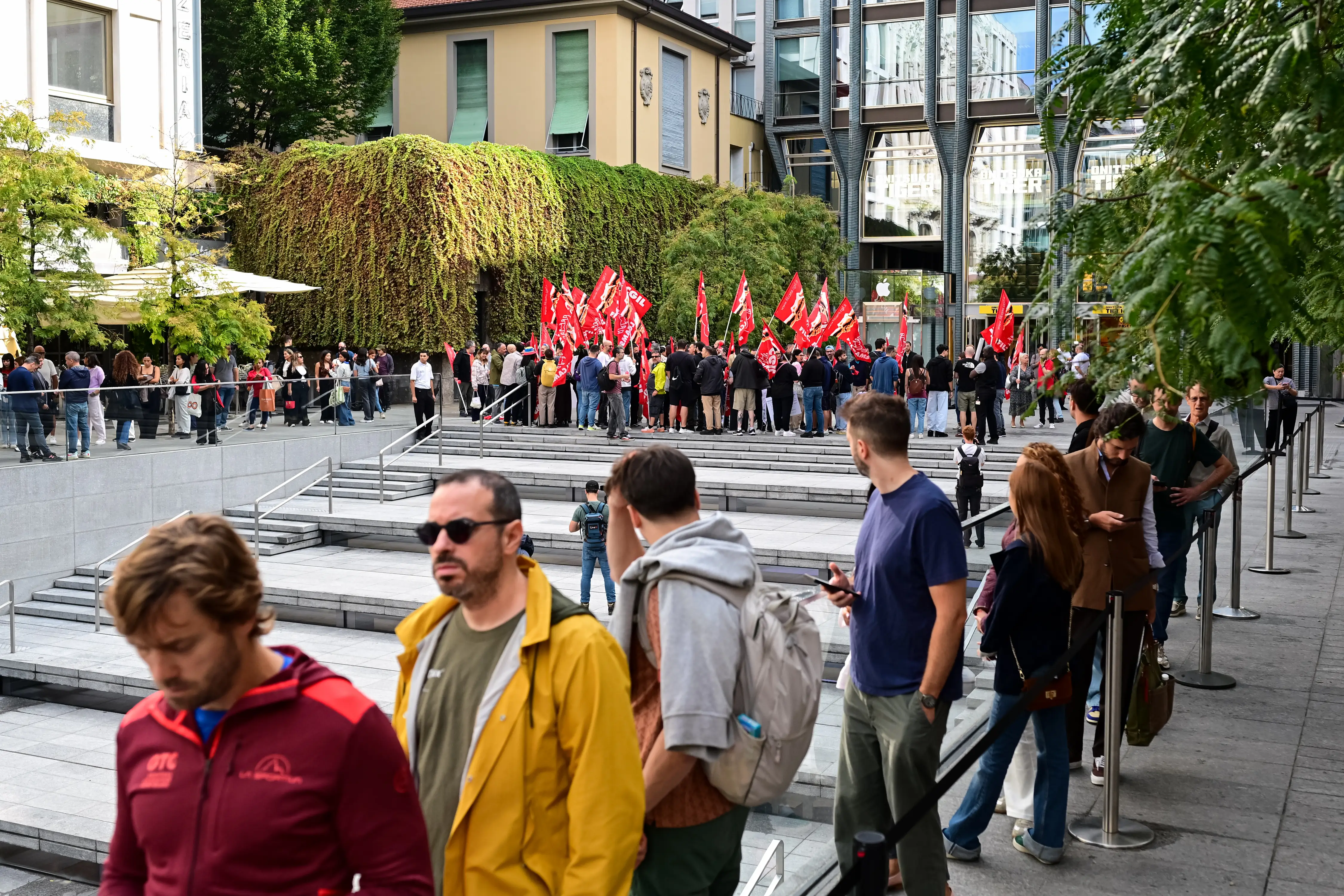 A line of shoppers outside the Milan Apple store. In the background, protesters gather.