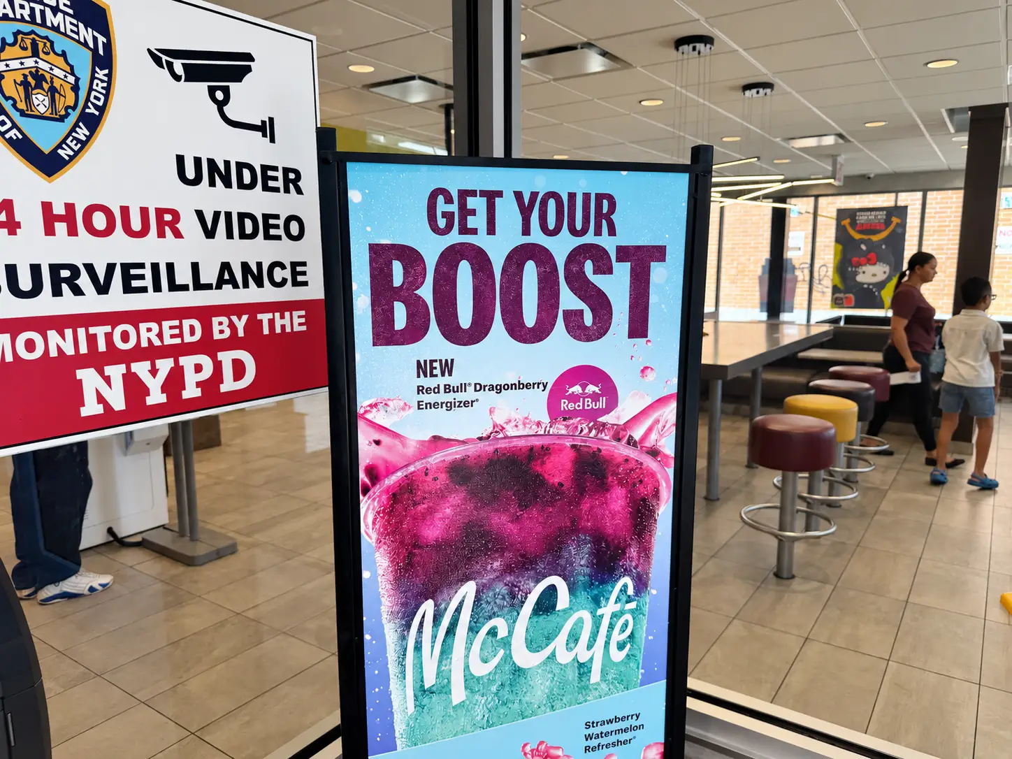McCafé digital ad for Red Bull Dragonberry drinks stands beside an NYPD surveillance sign inside a restaurant.