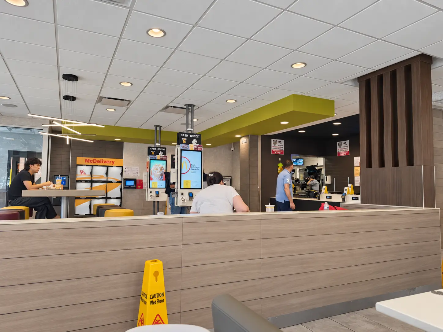 McDonald's restaurant interior with self-order kiosks, seating, counter area, and a yellow wet floor caution sign.