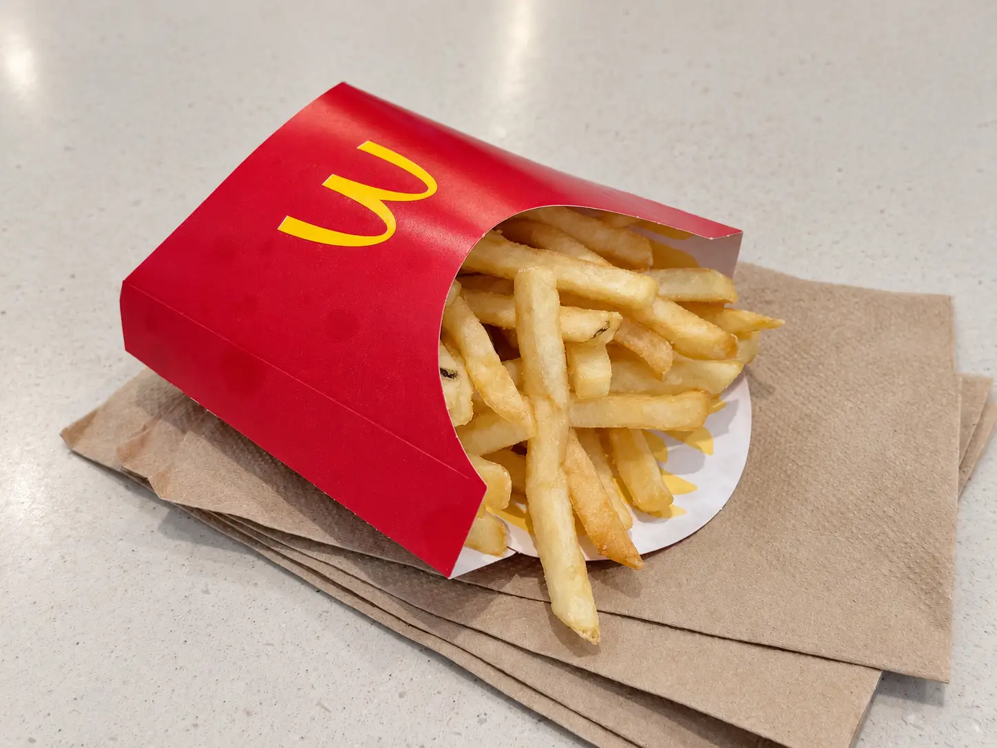 McDonald’s french fries in a red carton sit on brown napkins on a light countertop.