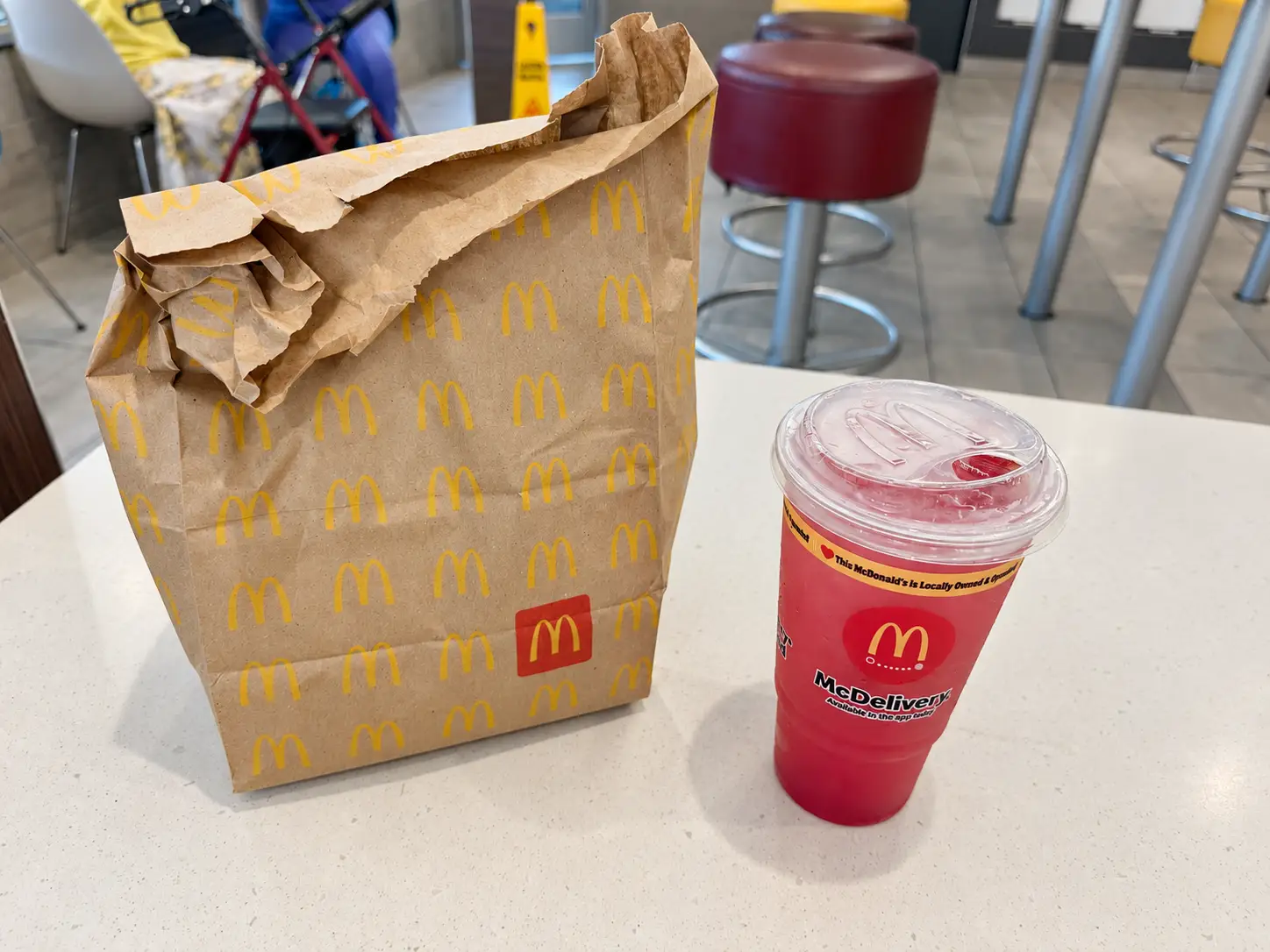 McDonald's paper takeout bag and red lidded drink sit on a restaurant table near stools.