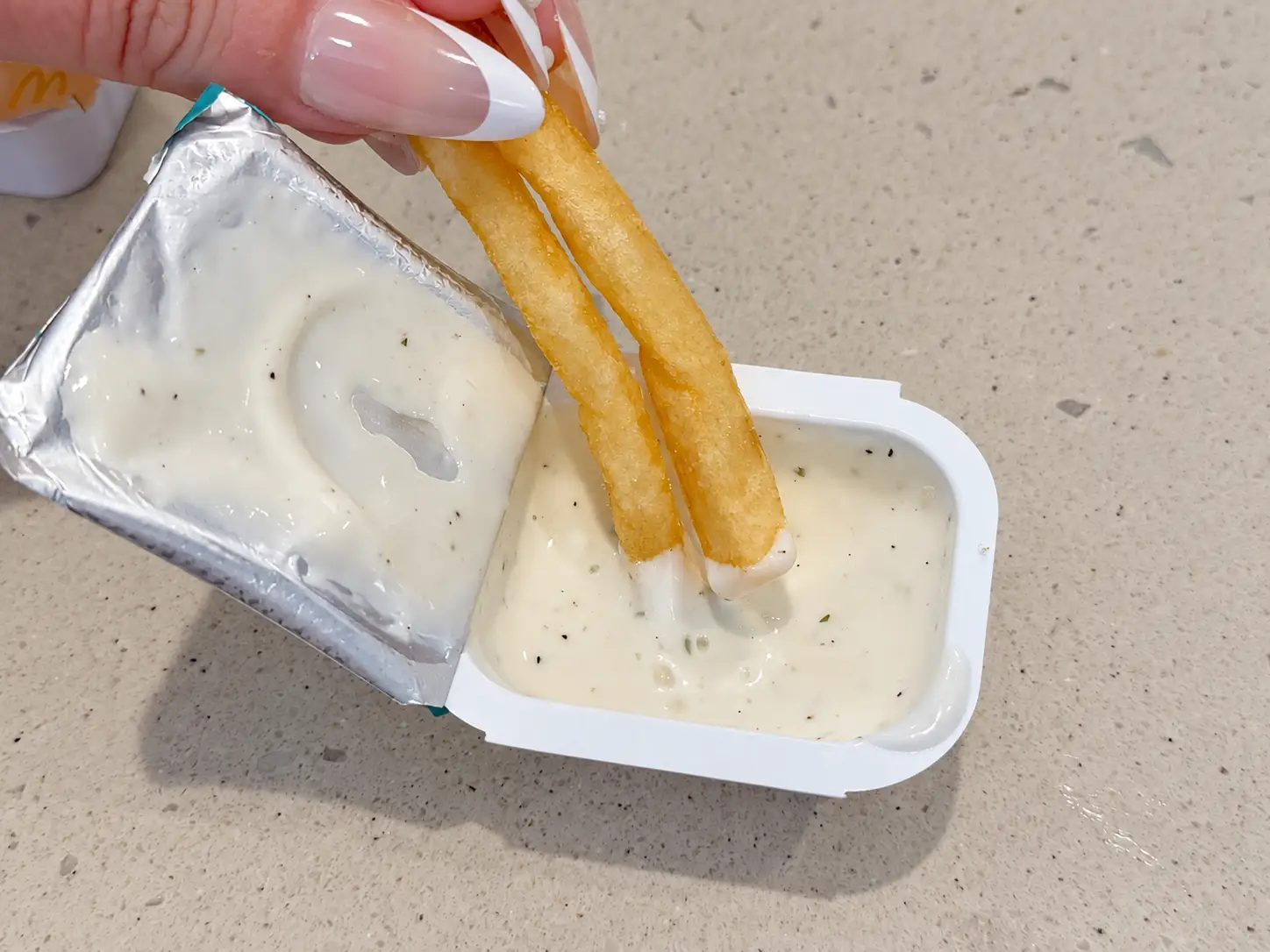Two french fries are dipped into a white creamy sauce packet on a speckled countertop.