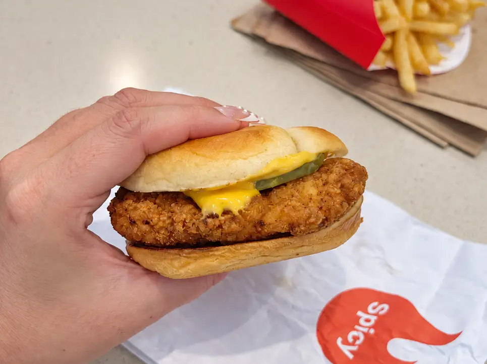 Hand holding a spicy chicken sandwich on McDonald's packaging with fries on a counter in the background.