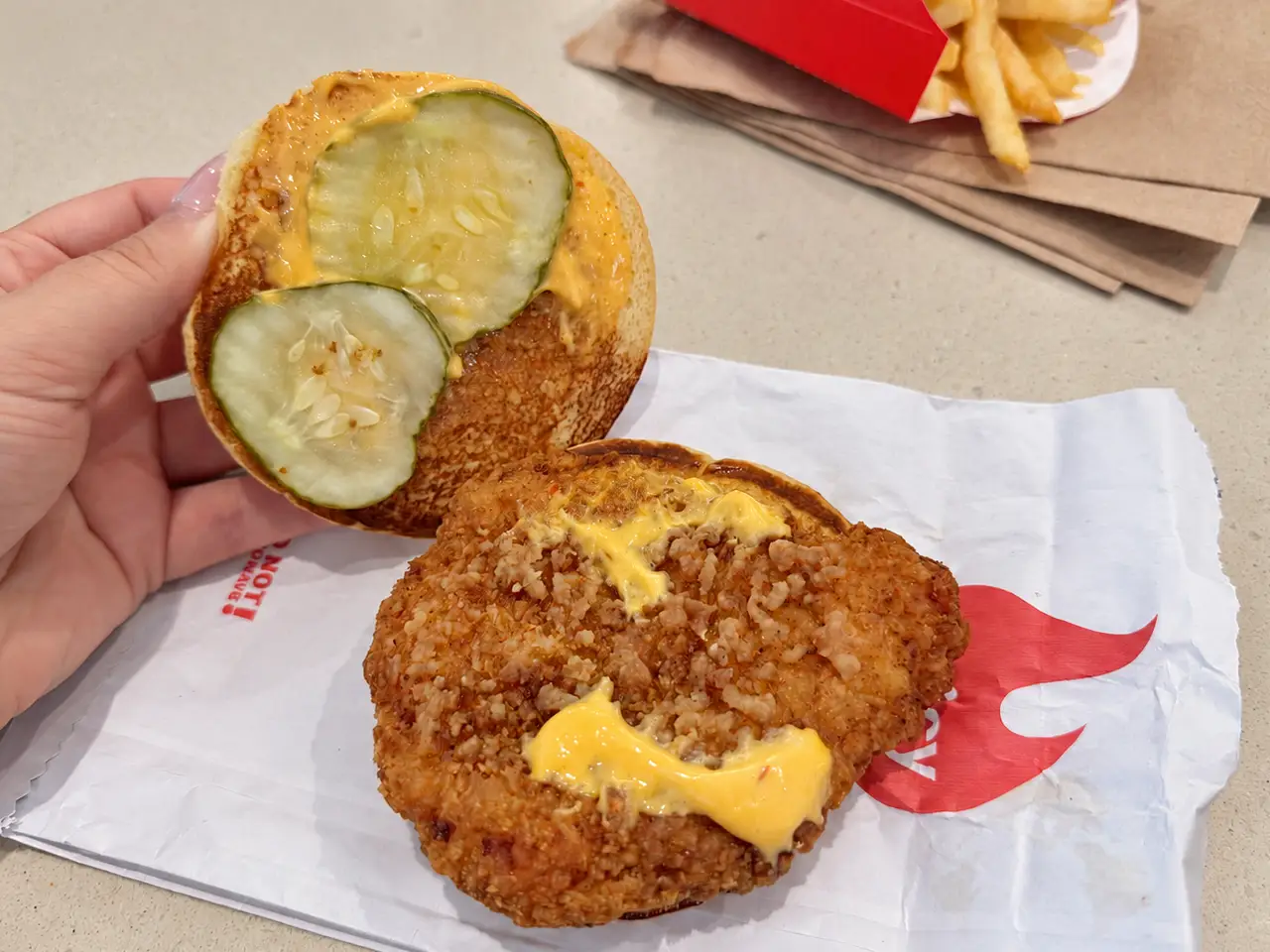 An open fried chicken sandwich with pickles and sauce sits on branded paper beside fries in a red carton.