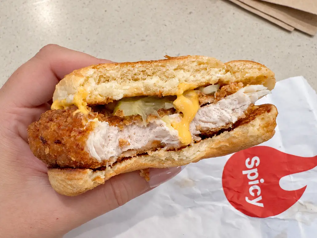 Hand holding a partially eaten spicy chicken sandwich over a wrapper, with fries and napkins on a countertop.