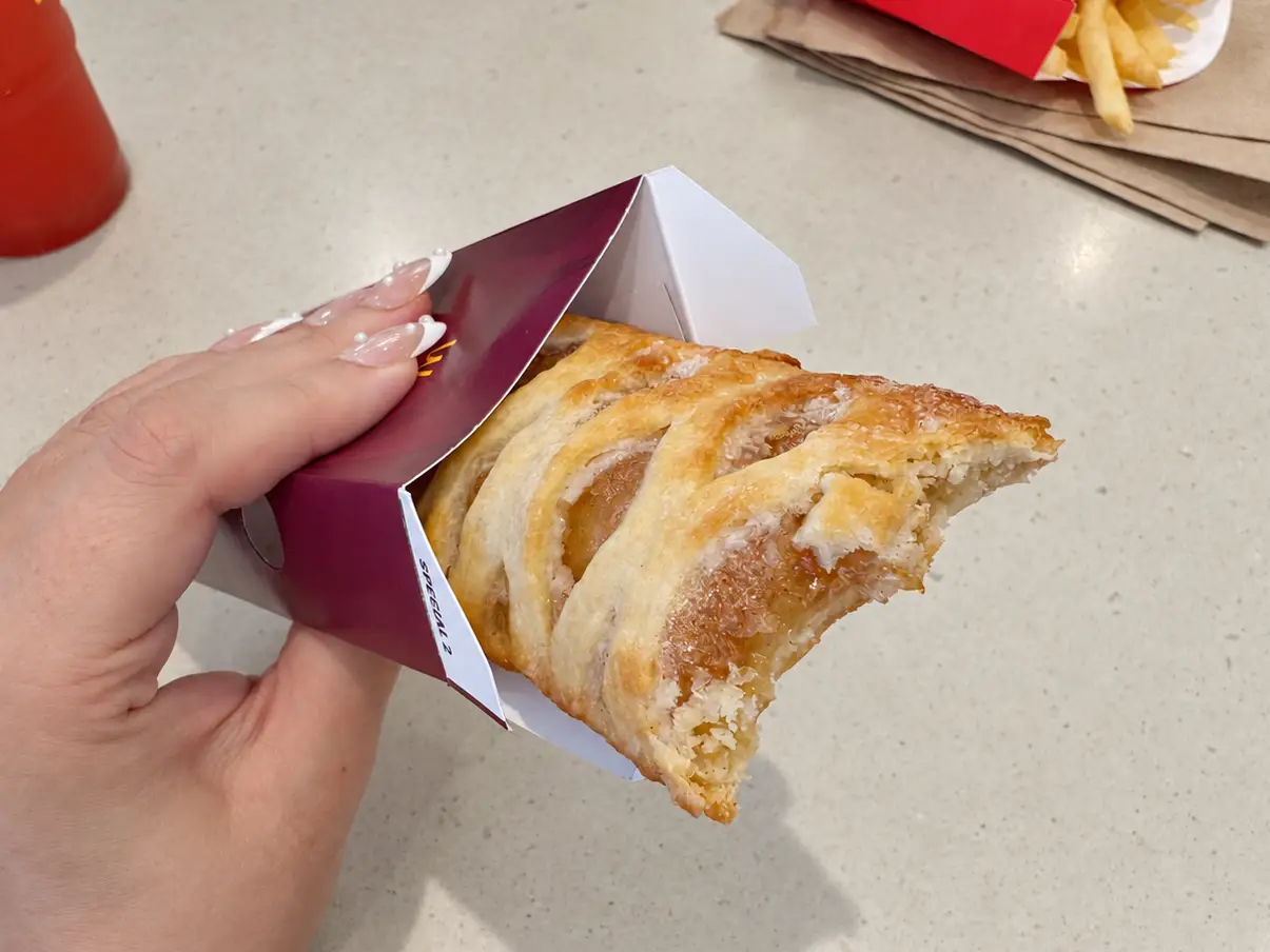 Hand holding a bitten pastry in a McDonald's box on a table with fries and a red drink nearby.