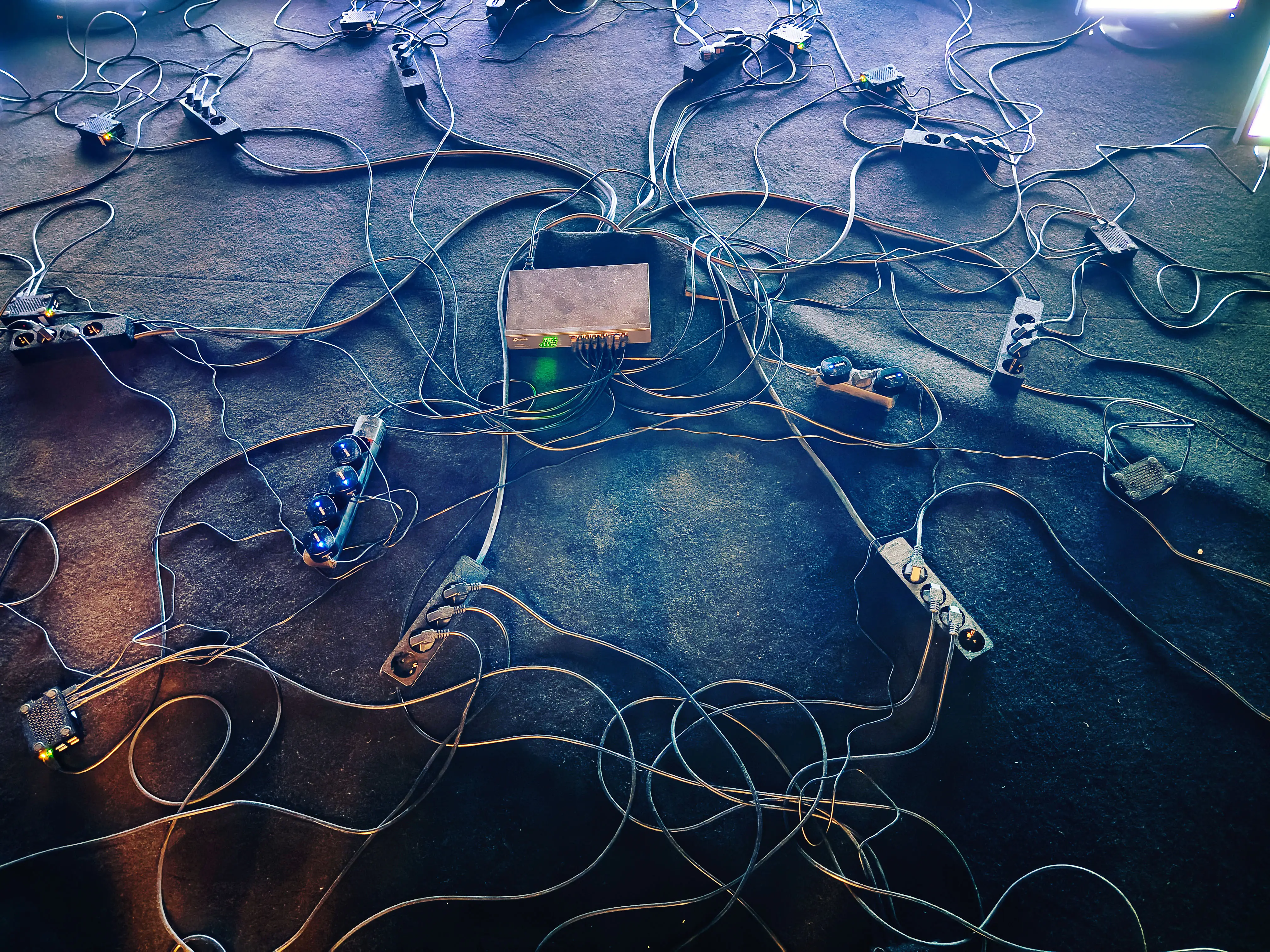 Network switches, power strips, adapters, and tangled cables cover a dark carpeted floor during equipment setup.