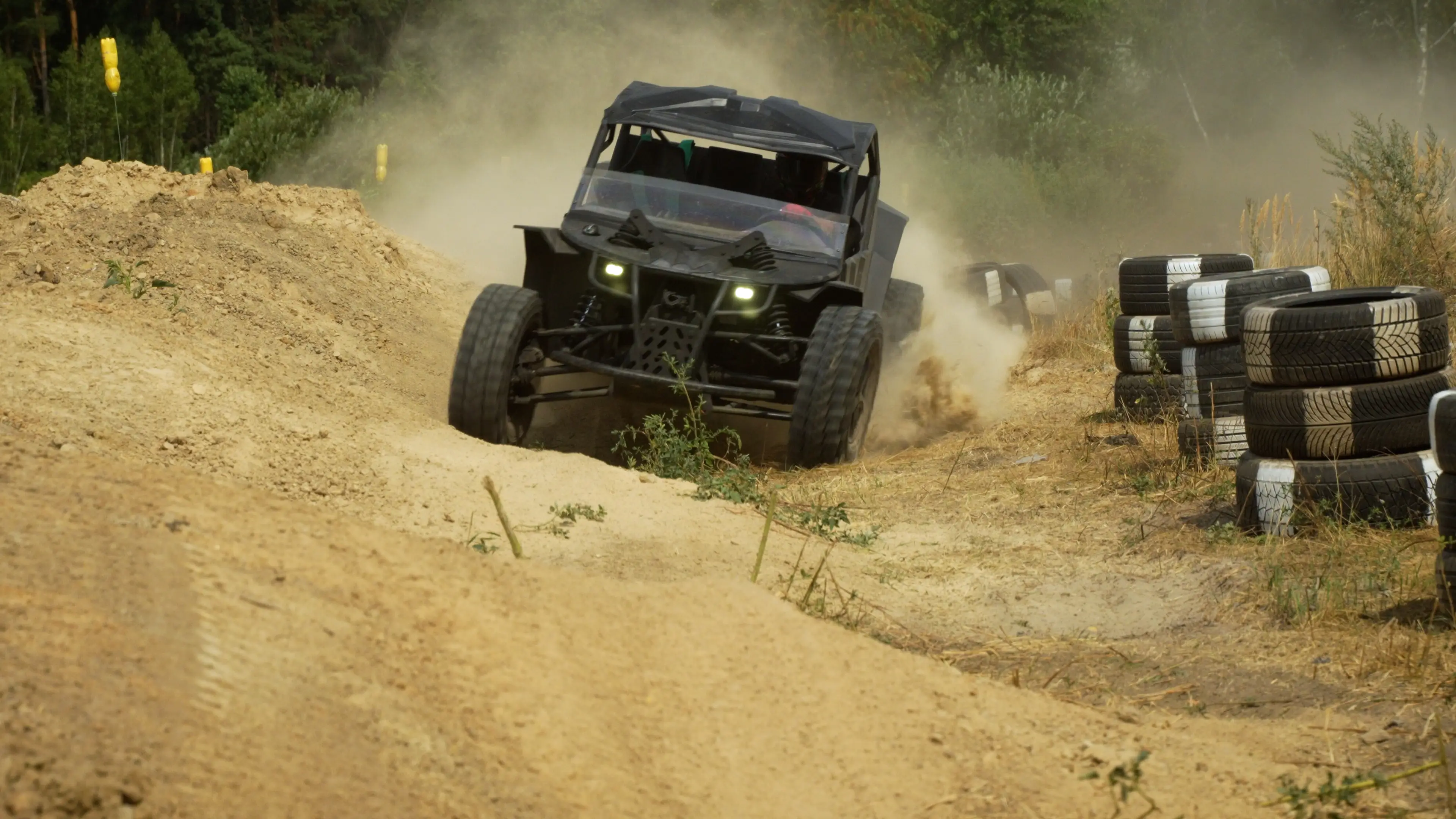 Off-road buggy drives through a dusty dirt track beside stacked tire barriers in a wooded outdoor course.