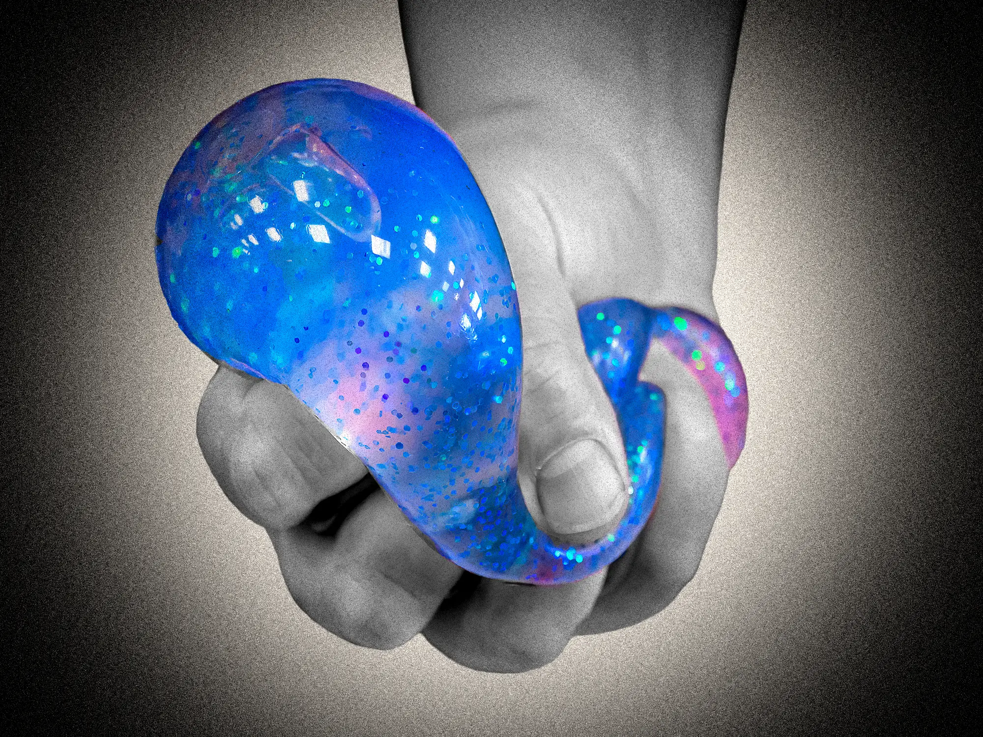 A hand holds a glossy blue and pink glittery flexible object against a grainy gray background.