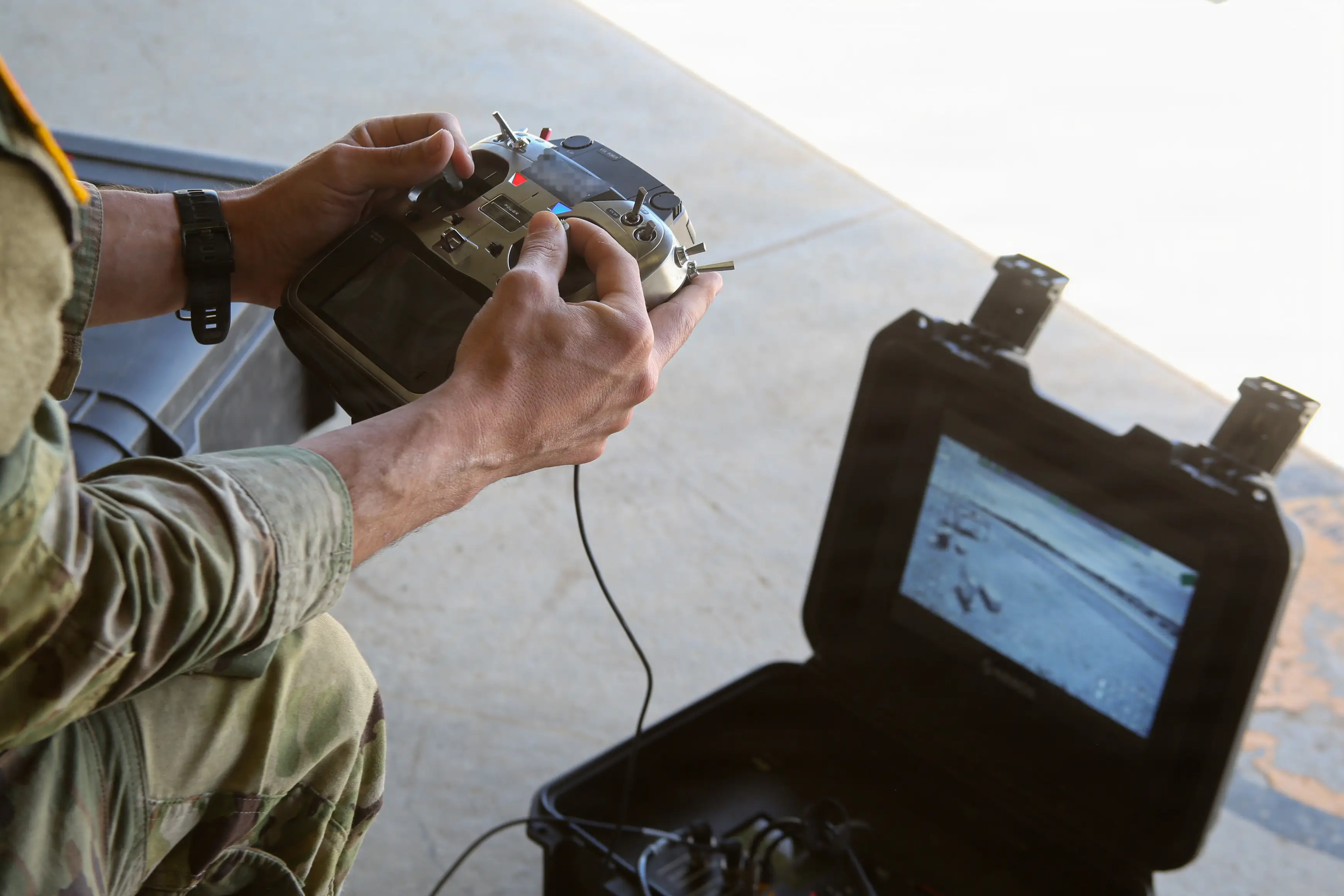 A US Army soldier holds a drone controller and crouches in front of a computer showing the drone's camera feed.
