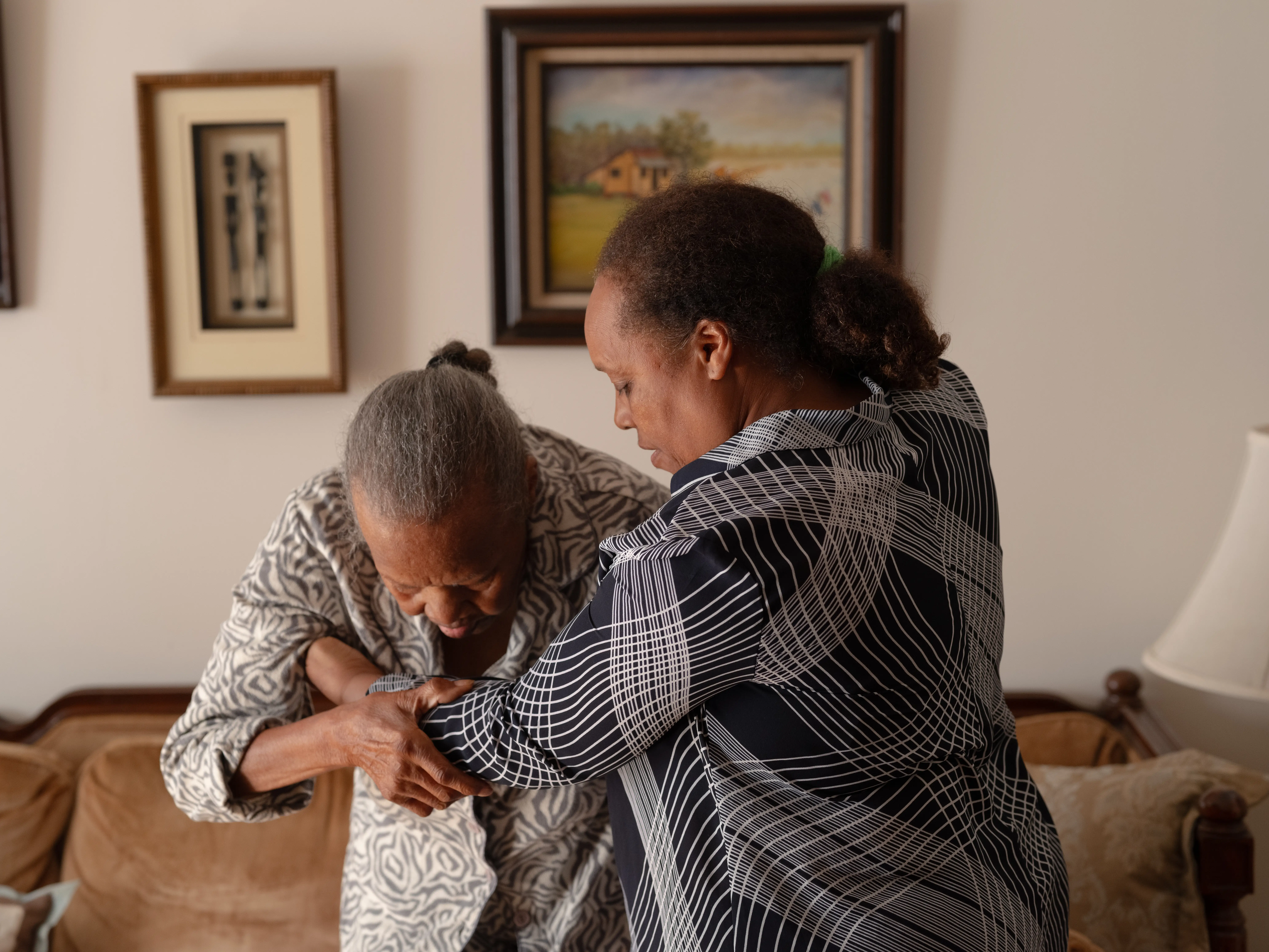 Two people stand beside a couch, with one supporting the other's arm in a living room.