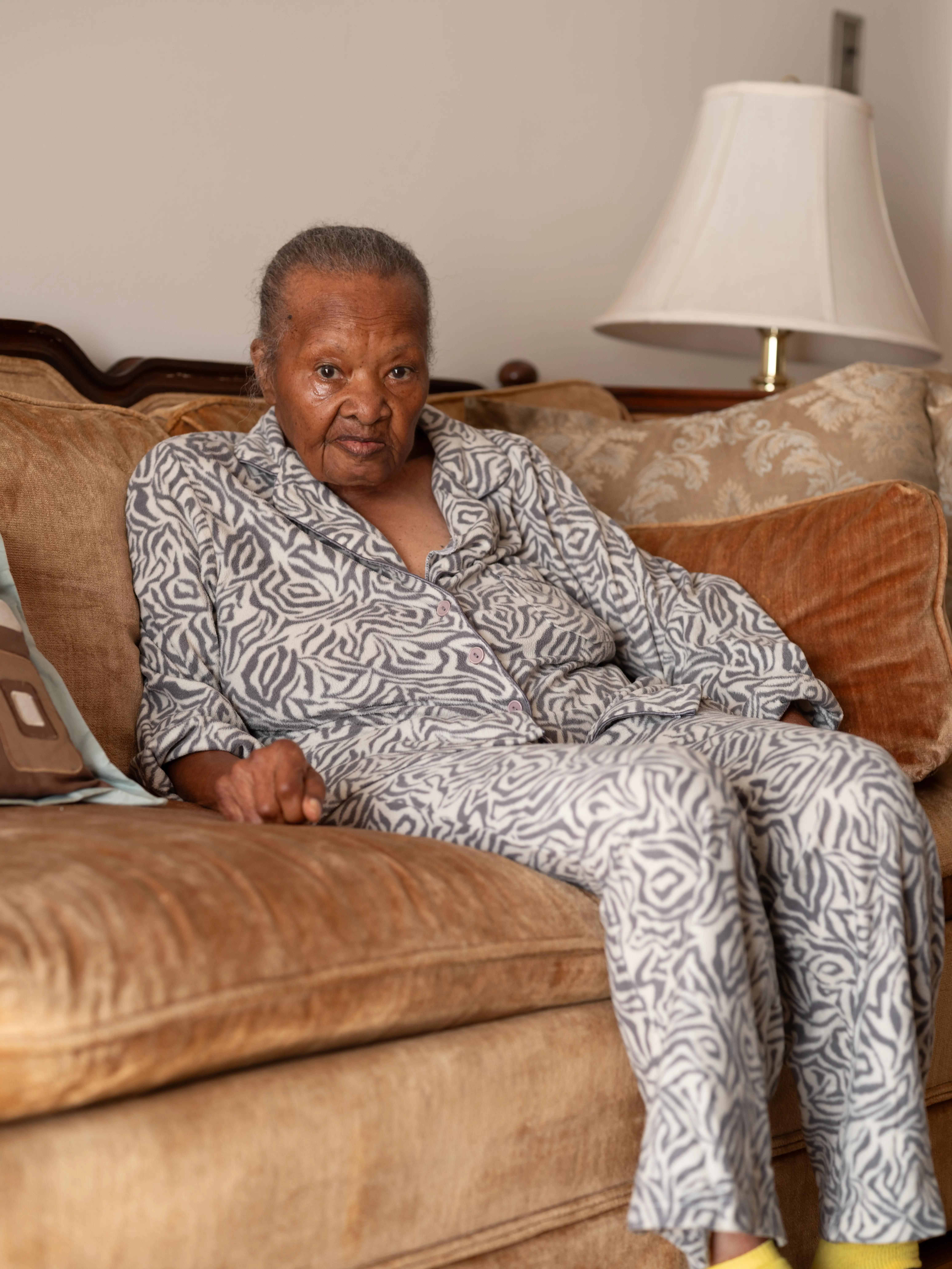 A person in patterned pajamas sits on a tan couch with pillows and a table lamp nearby.