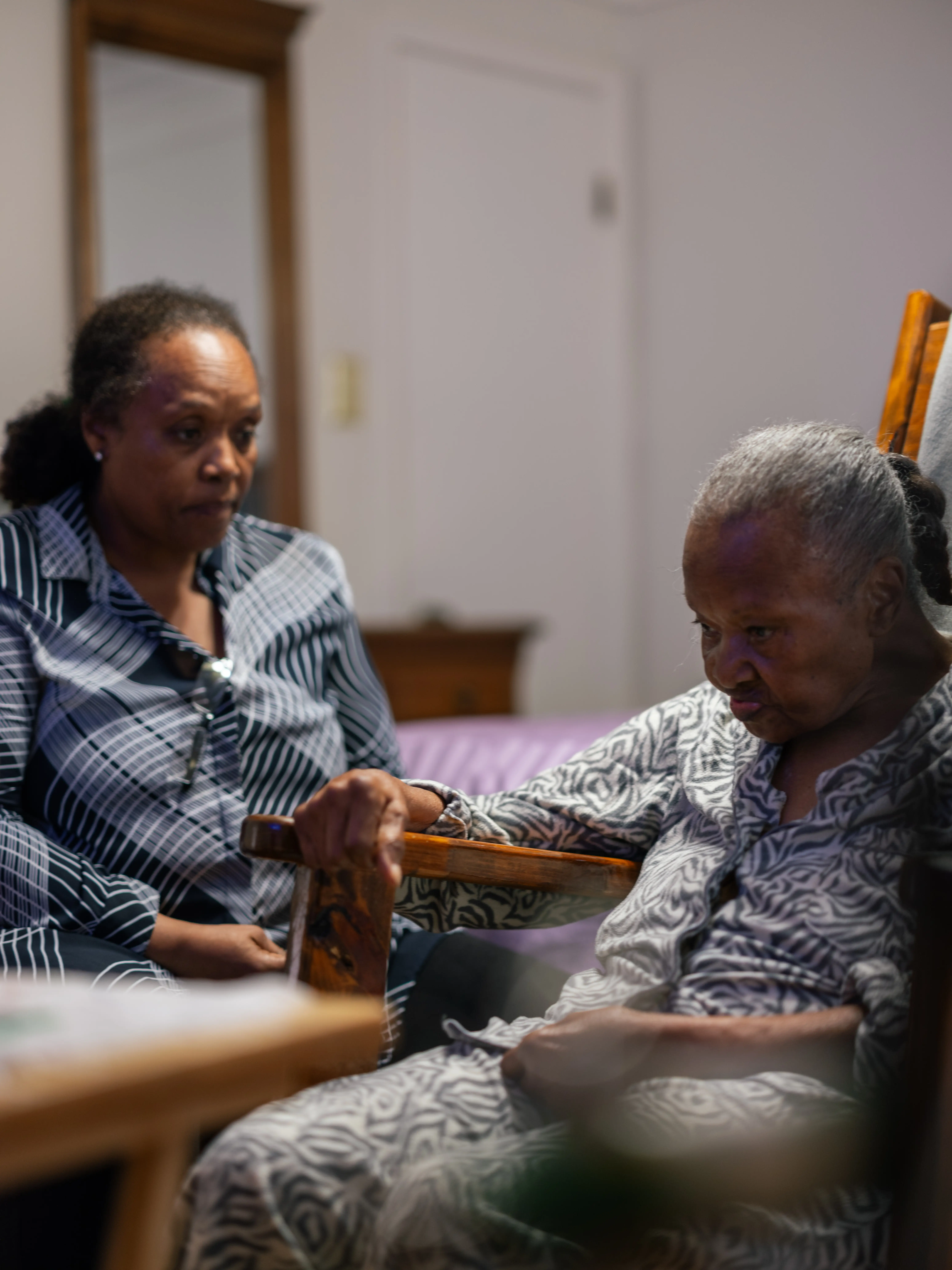 Two people sit in a bedroom, with one resting a hand on a wooden chair arm.