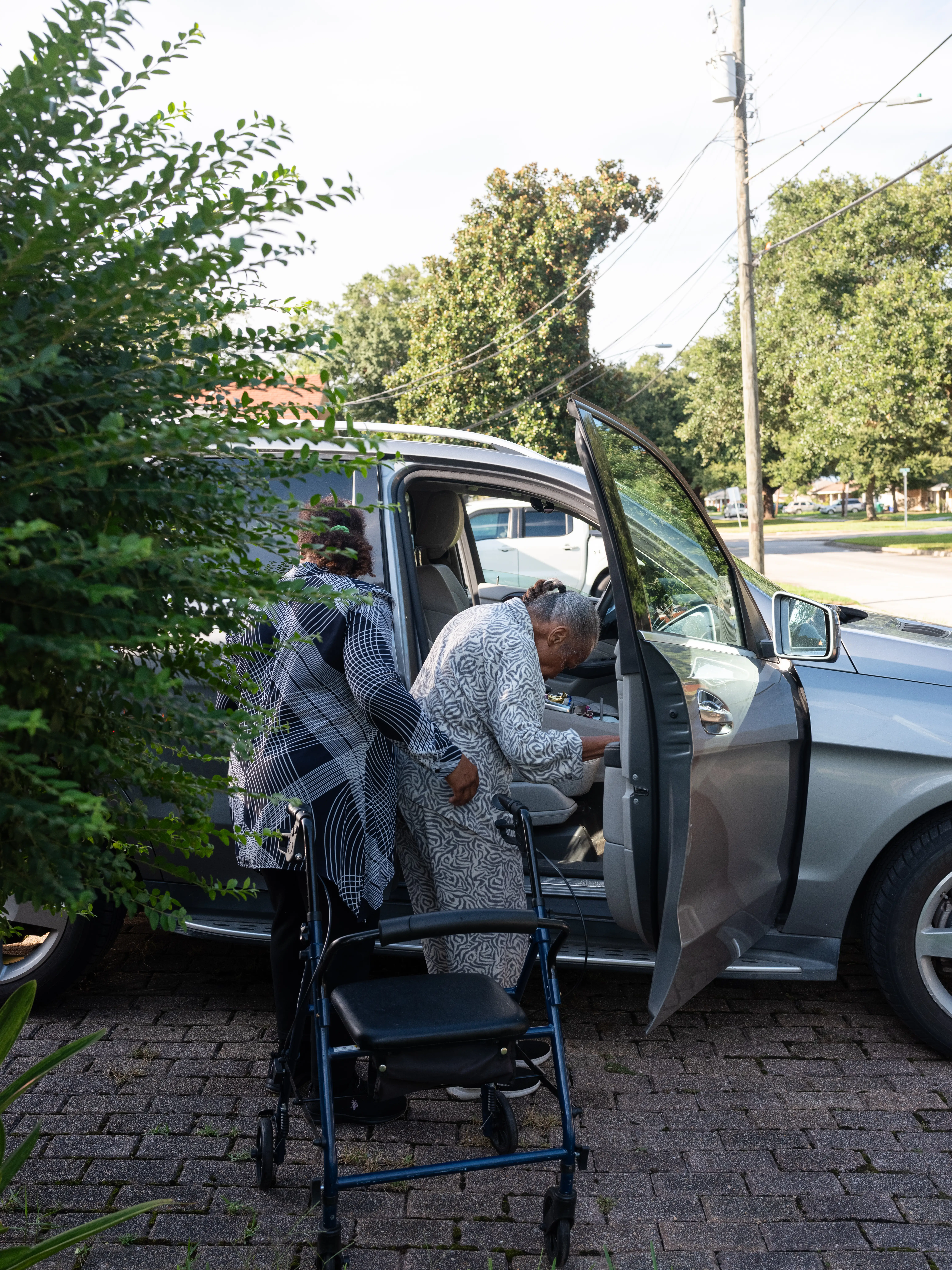 Two people stand beside an open car door with a rolling walker on a residential driveway.