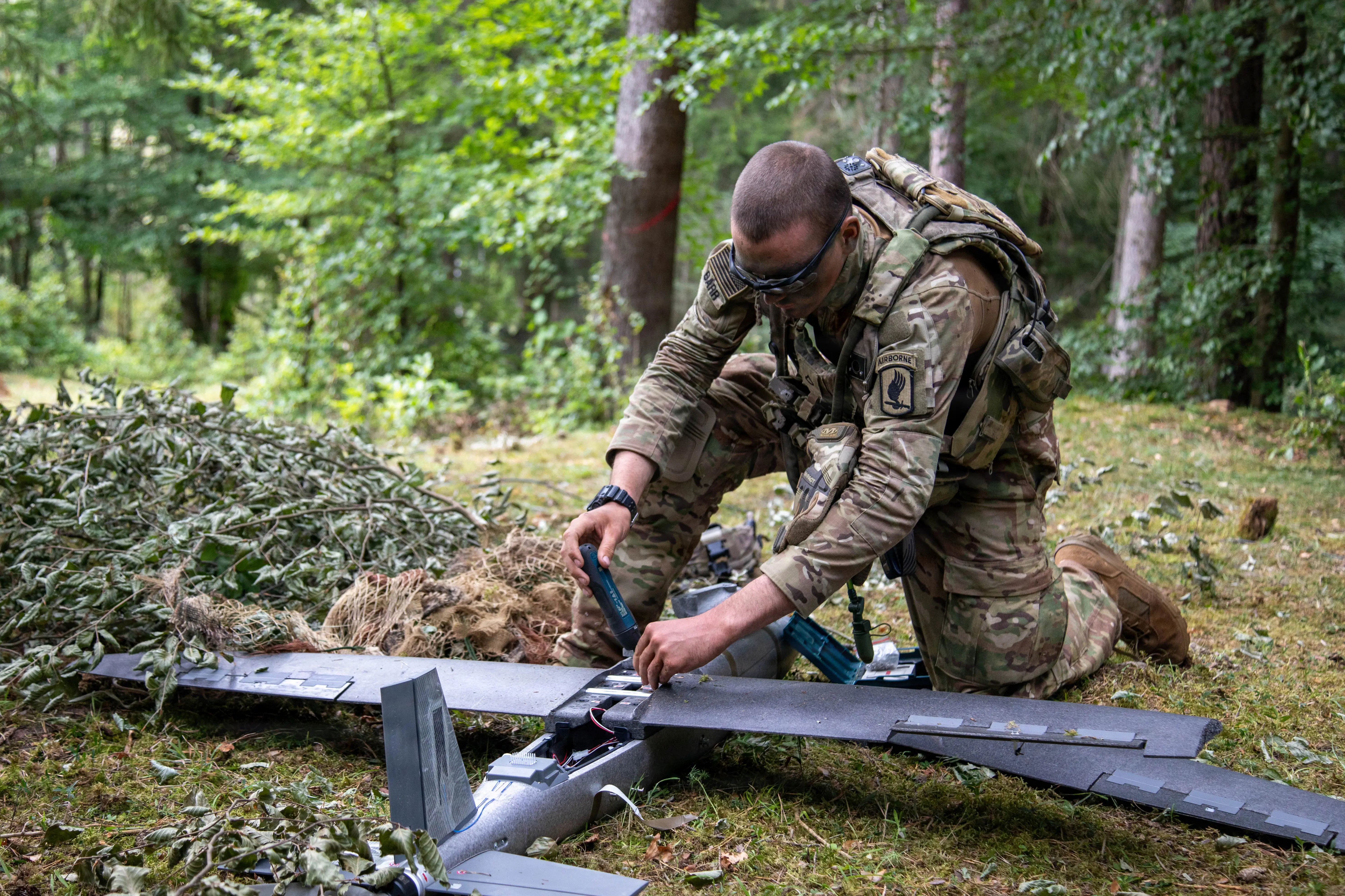 A US Army soldier conducts maintenance on a large fixed-wing drone in a forest.