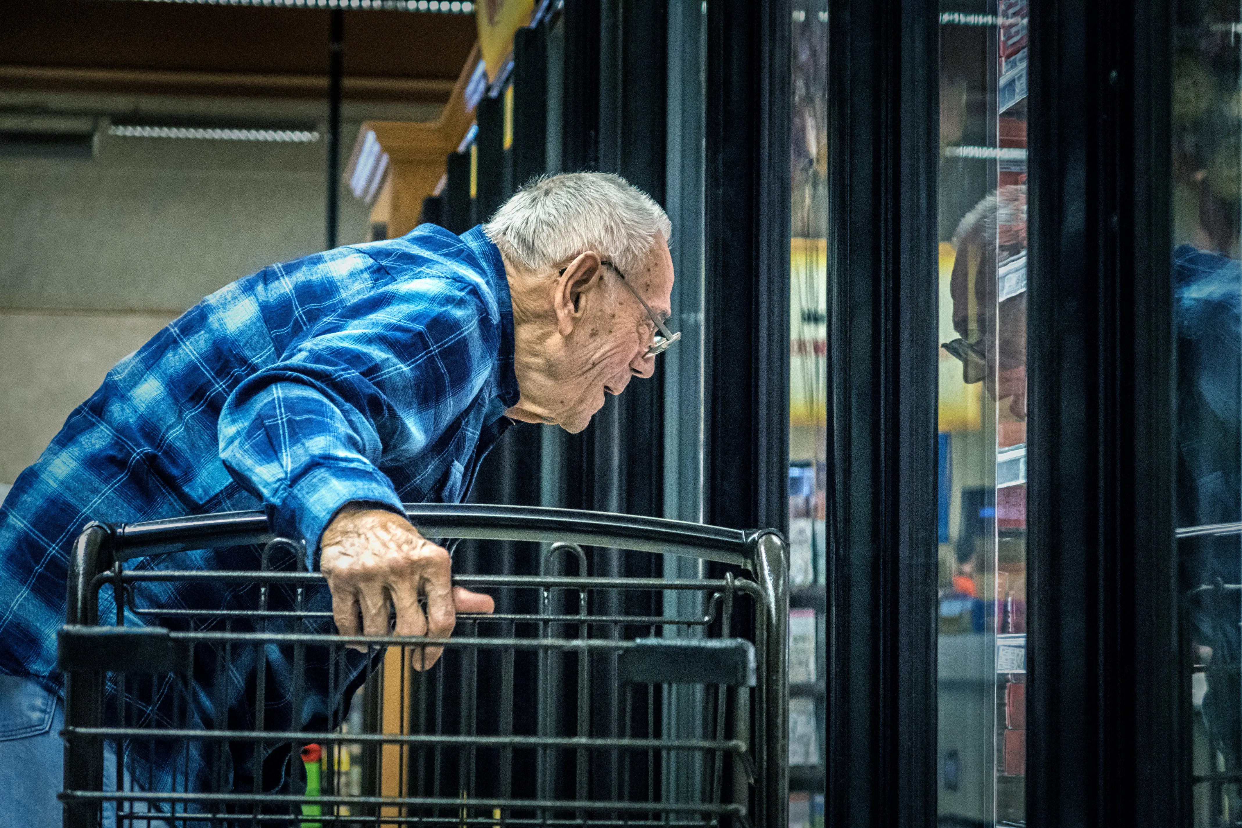 Shopper in a blue plaid shirt leans over a cart to look into refrigerated grocery cases.