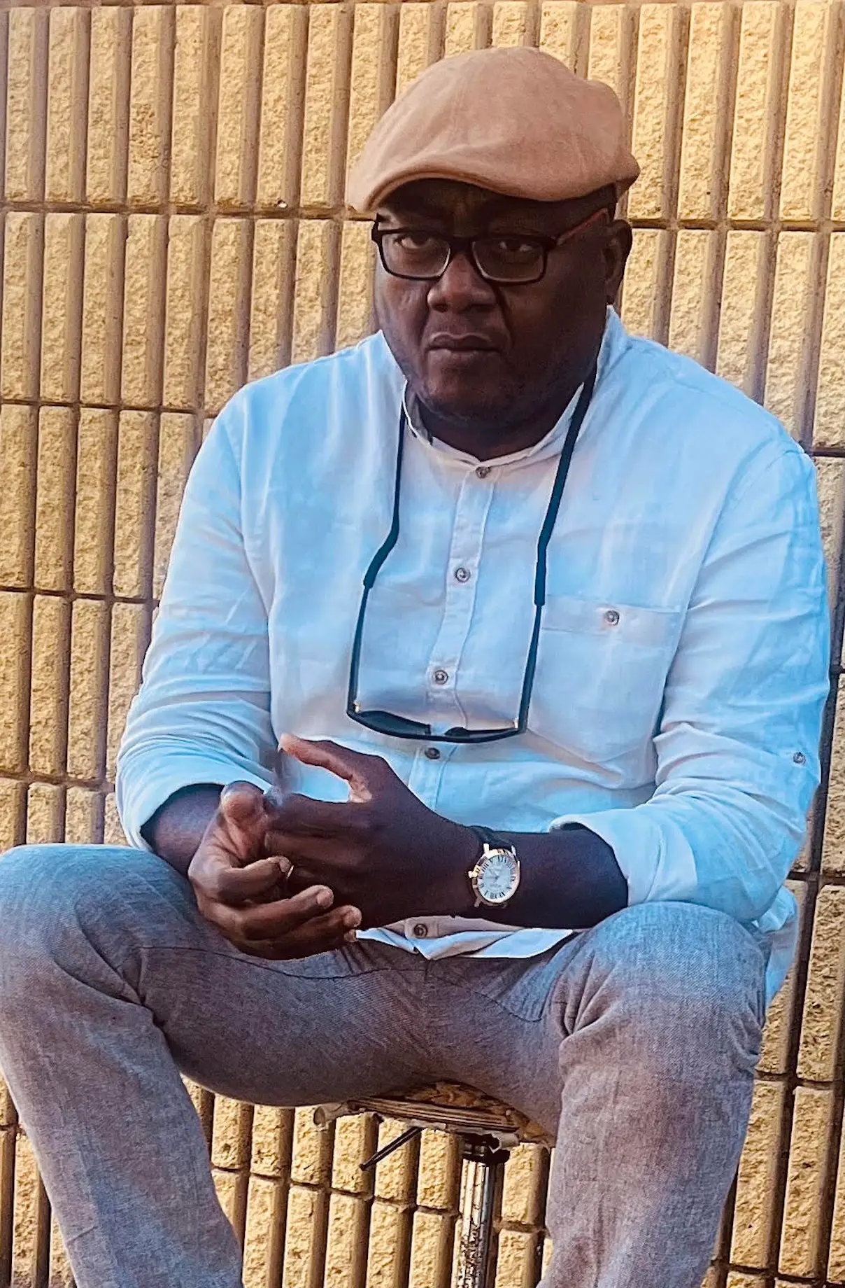 Osahon Aburime, a former Lyft driver, sits with sunglasses around his neck with a tan wall behind him, wearing a tan cap.