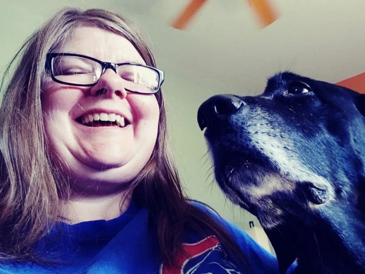 A woman in a blue sweatshirt is laughing with a dog beside her.