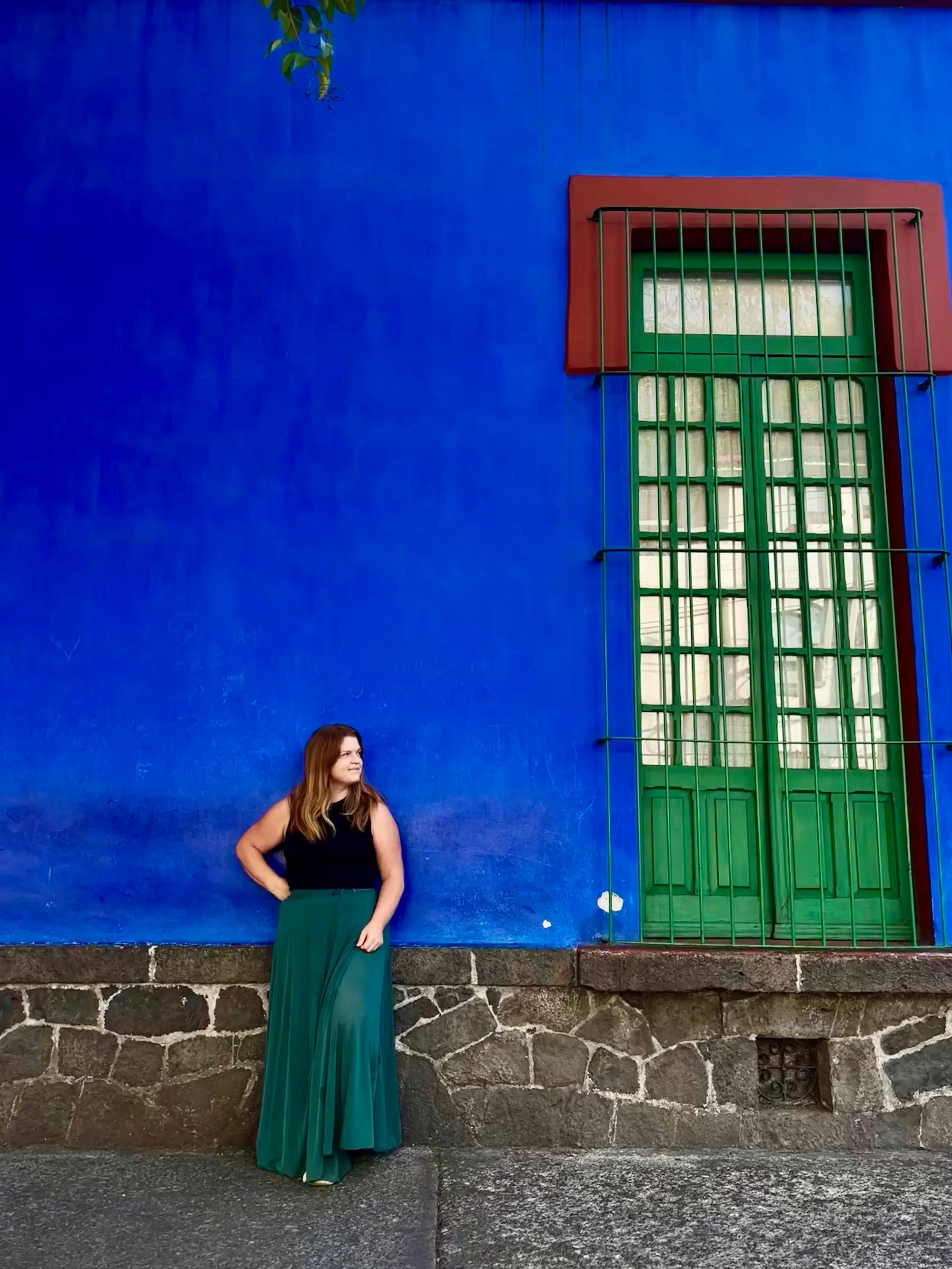 Person stands against a vivid blue wall beside a tall green barred window and stone base.