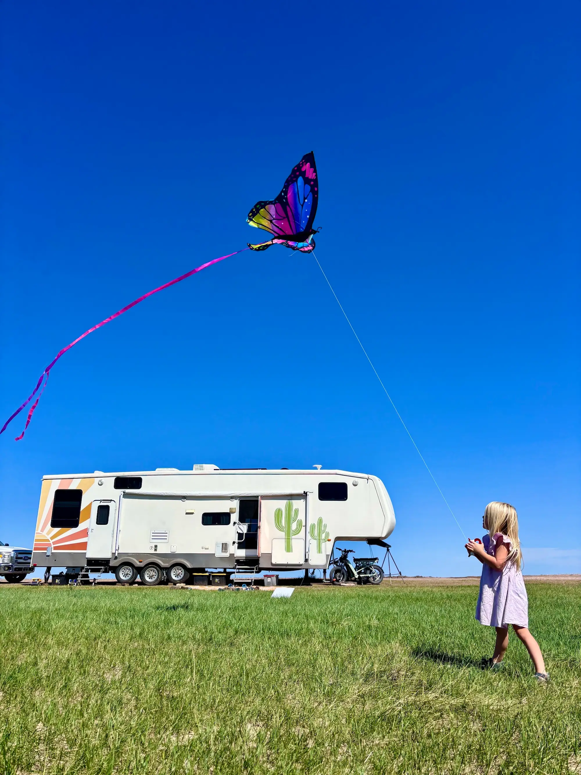 A person flies a colorful butterfly kite beside a camper trailer in a sunny grassy field.
