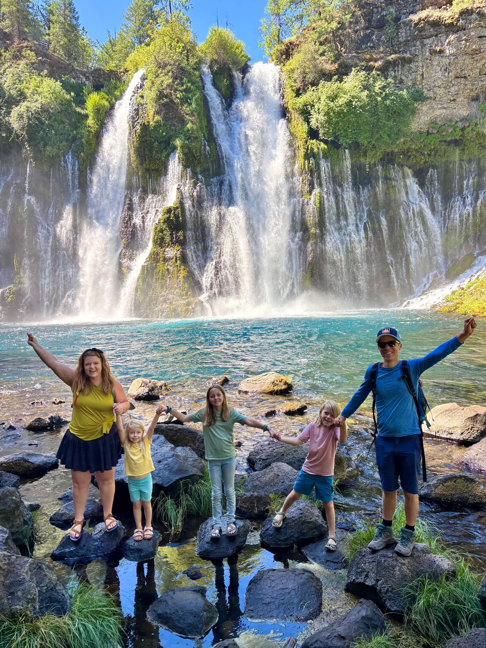 Five hikers pose on rocks beside a turquoise pool below a wide forested waterfall.