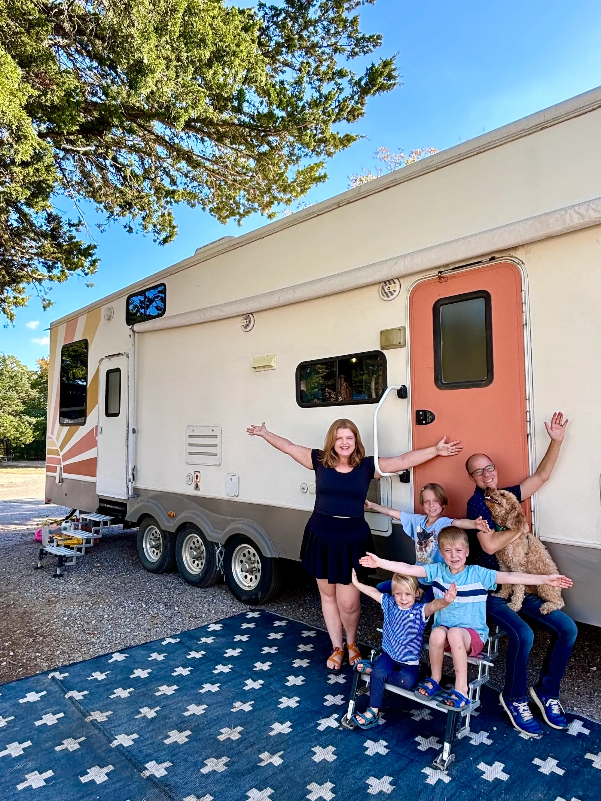Five people and a dog pose with raised arms outside a large travel trailer on a patterned outdoor rug.