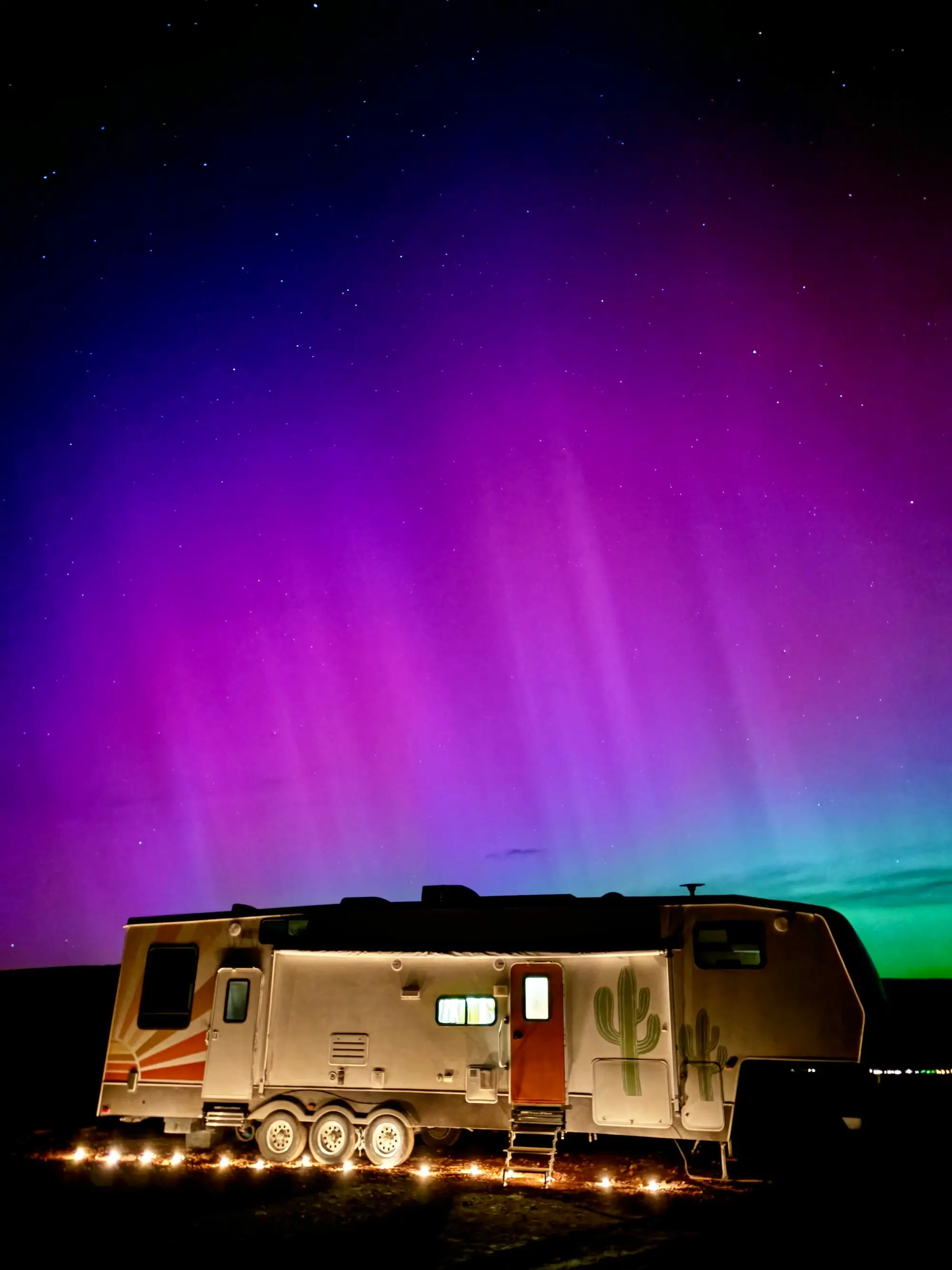 Illuminated travel trailer sits under a vivid purple and green aurora-filled night sky.