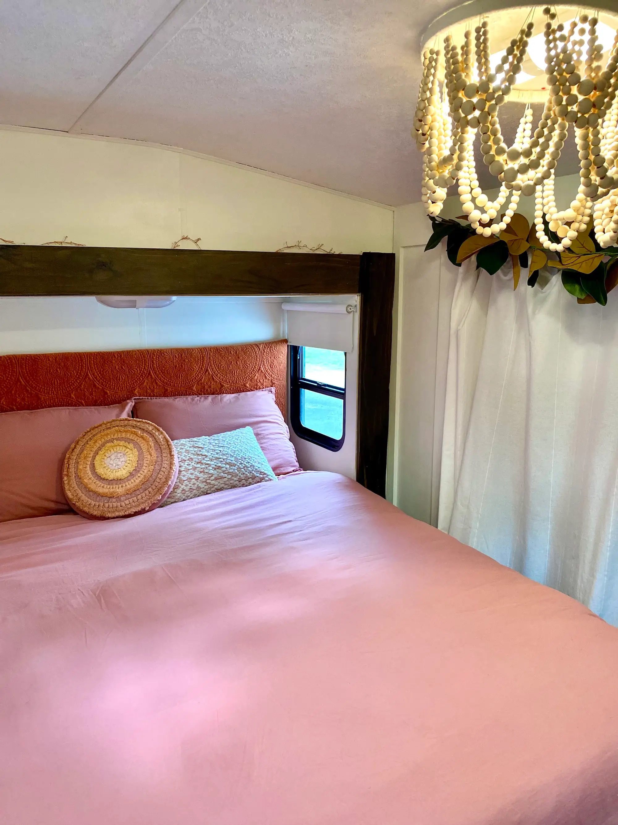 Cozy bedroom interior with pink bedding, decorative pillows, beaded ceiling light, curtains, and a small window.