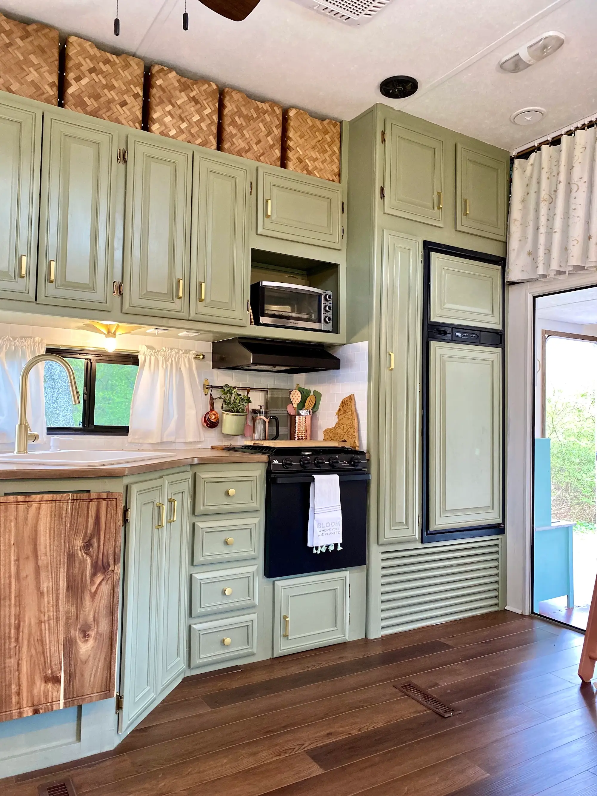 Compact kitchen interior with sage green cabinets, black stove, microwave, sink, refrigerator, woven baskets, and wood flooring.