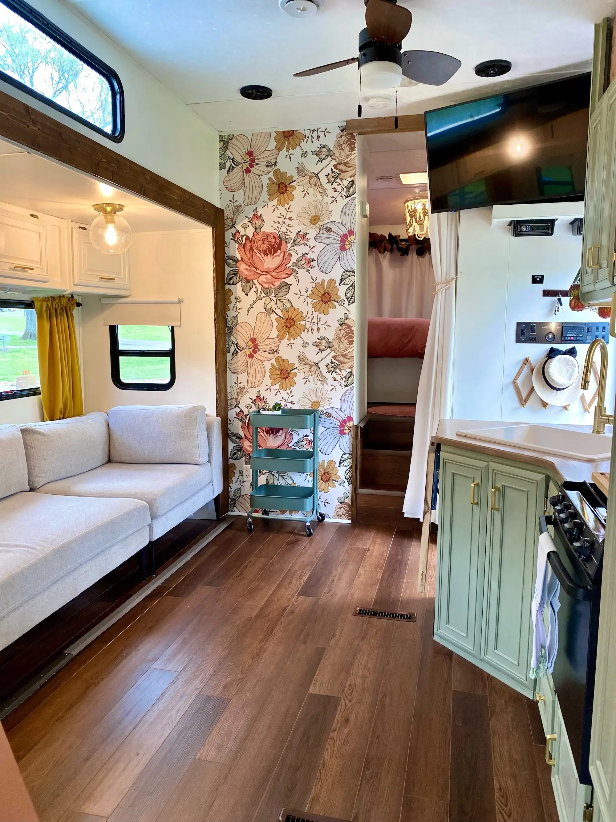 Renovated camper interior with a sofa, floral wallpaper, kitchenette, wall-mounted TV, and loft sleeping area.