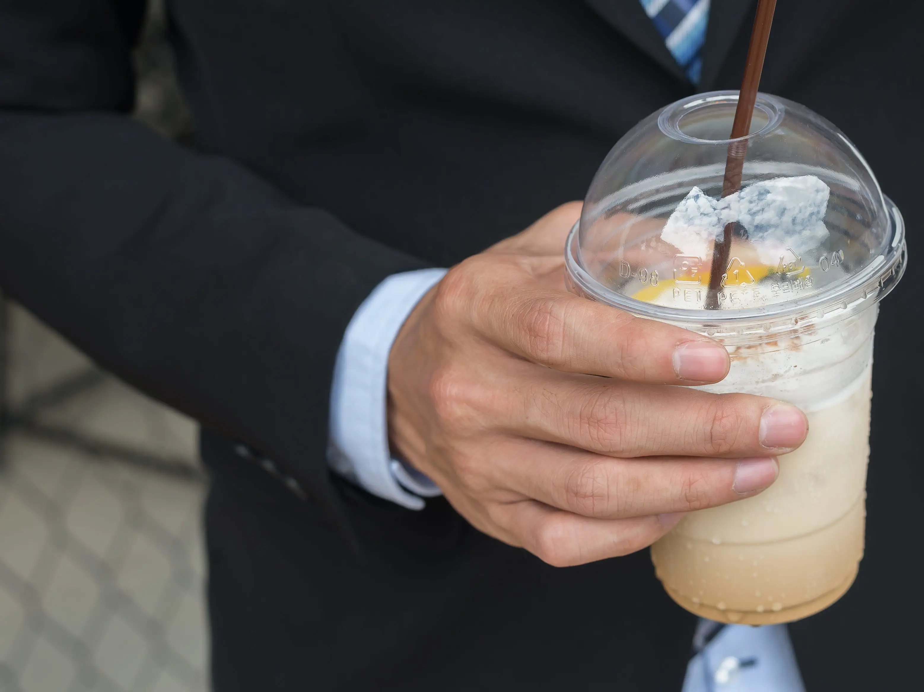 Close up of a person in a dark suit holding a clear plastic cup with an iced drink and straw.