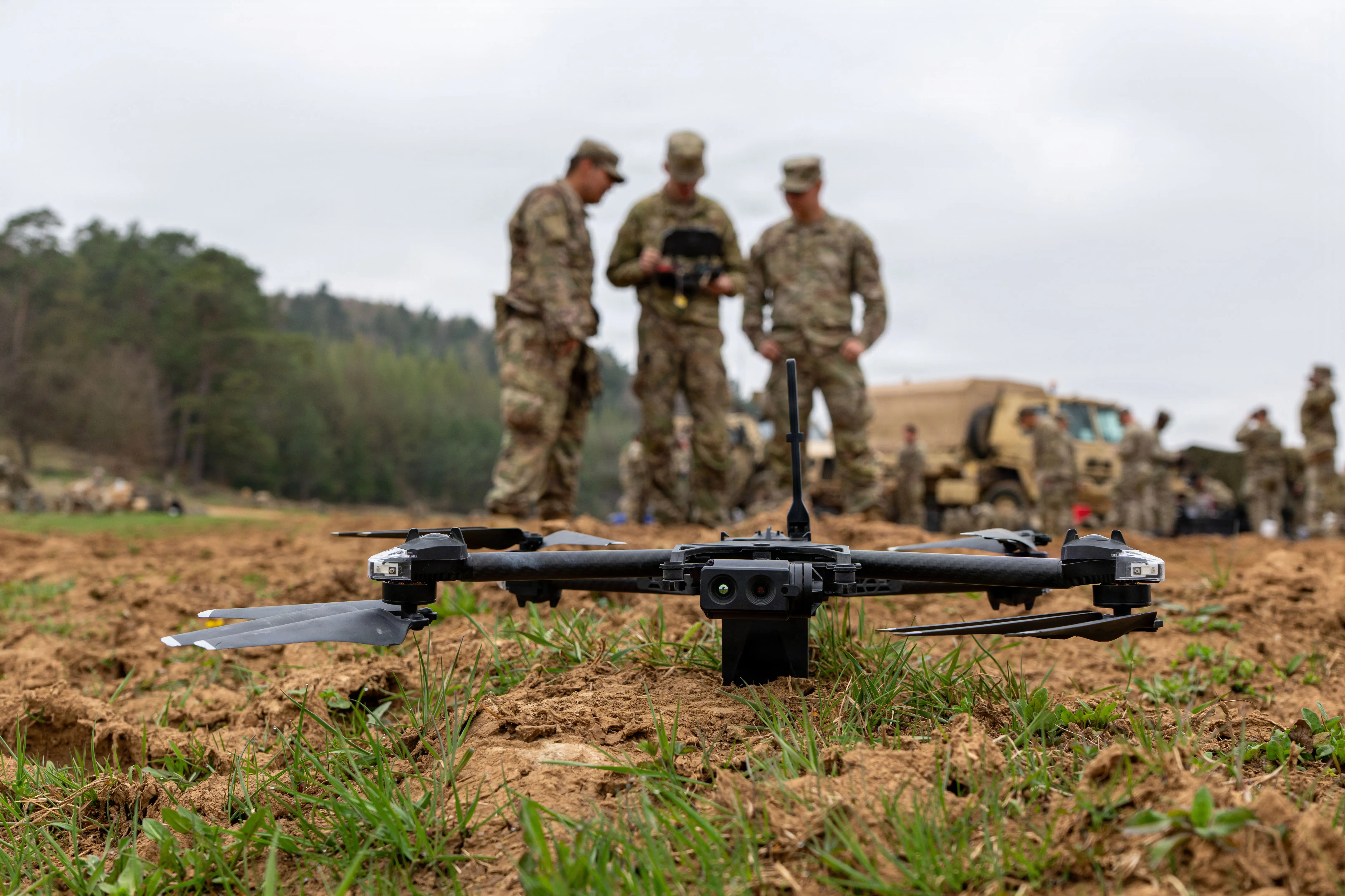US Army soldiers stand behind a small quadcopter drone that's sitting on the ground.