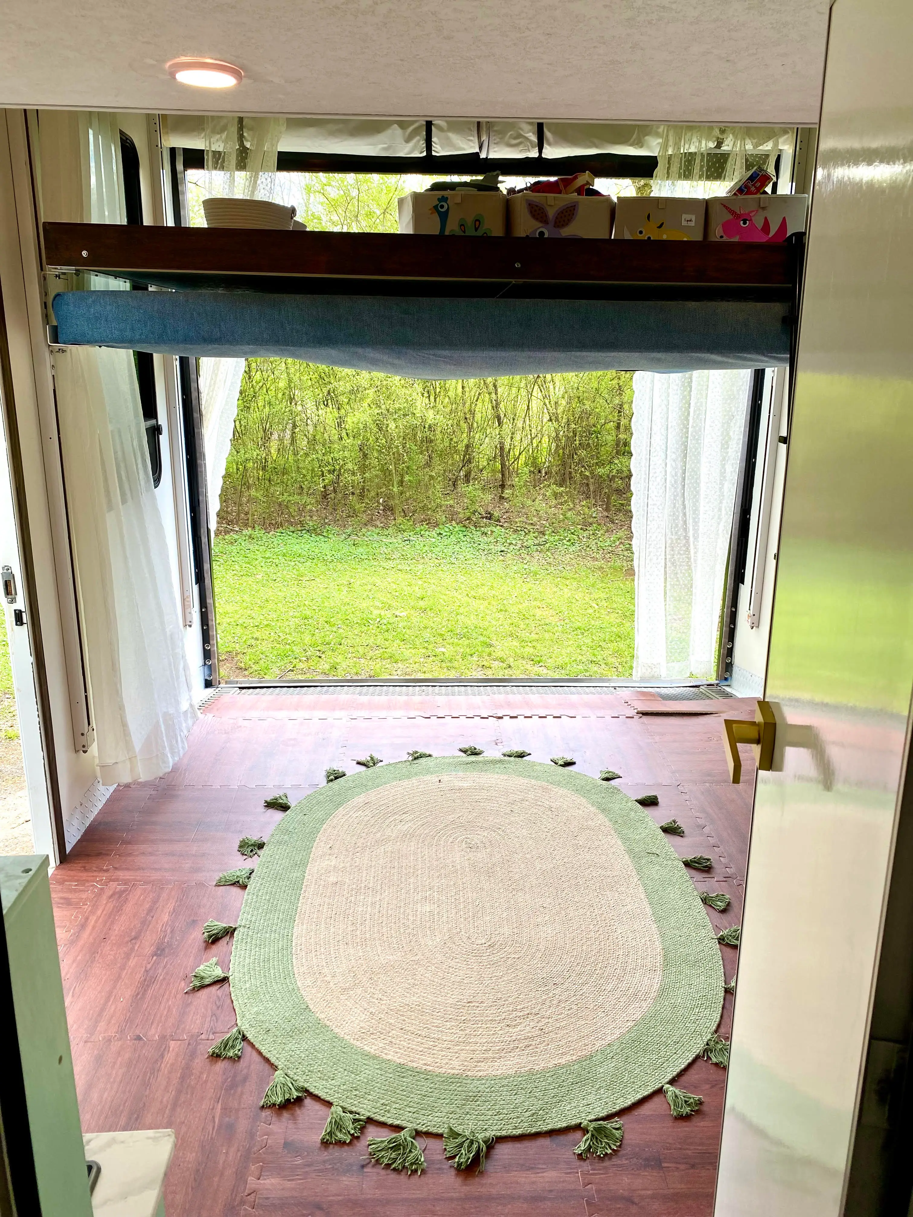 Compact interior with an open rear doorway, oval tasseled rug, curtains, shelf bins, and greenery outside.