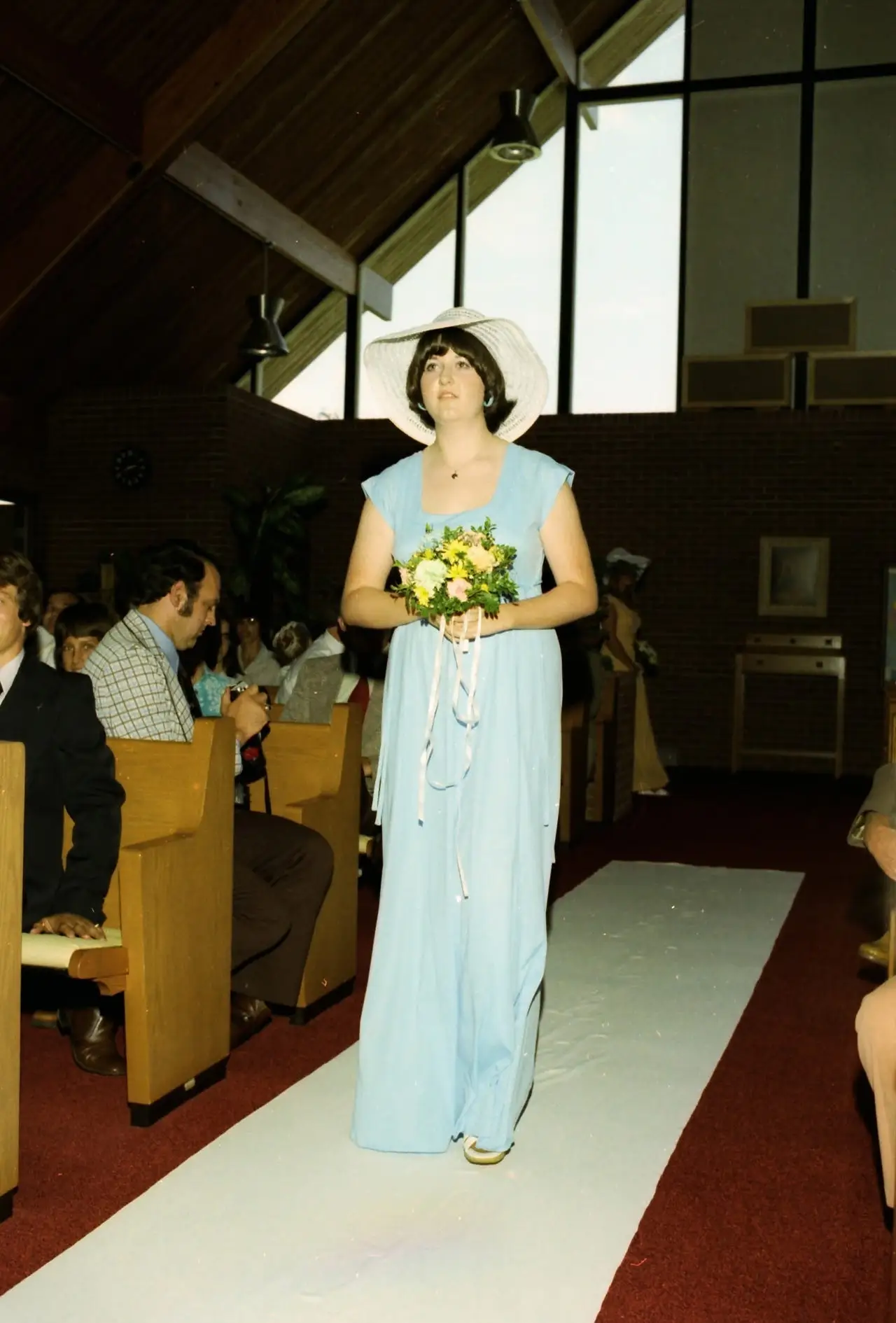 the author wearing a blue bridesmaid dress and white hat walking down the aisle at her friend's wedding