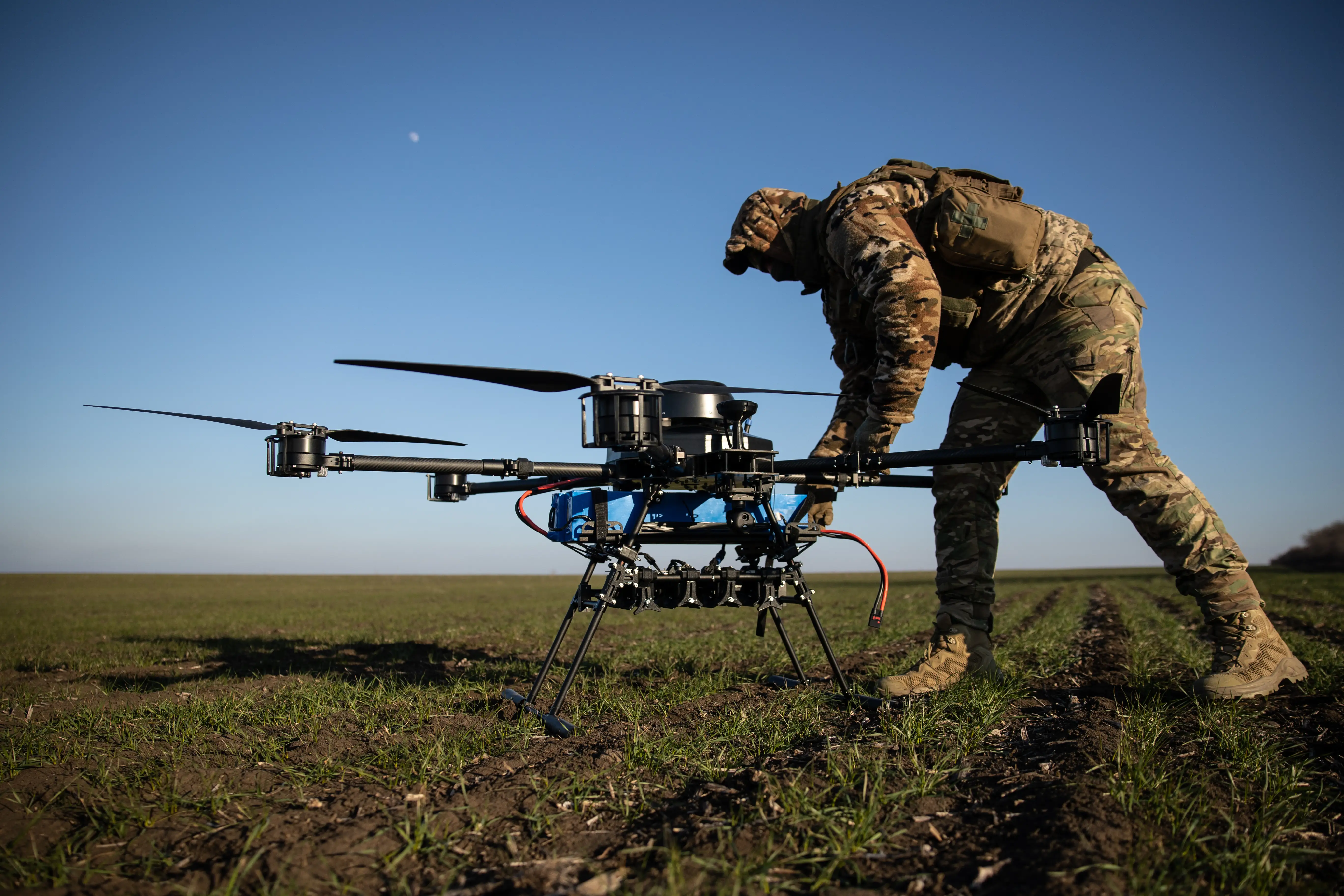 A camouflage-clad person adjusts a large multi-rotor drone in an open field under a clear sky.