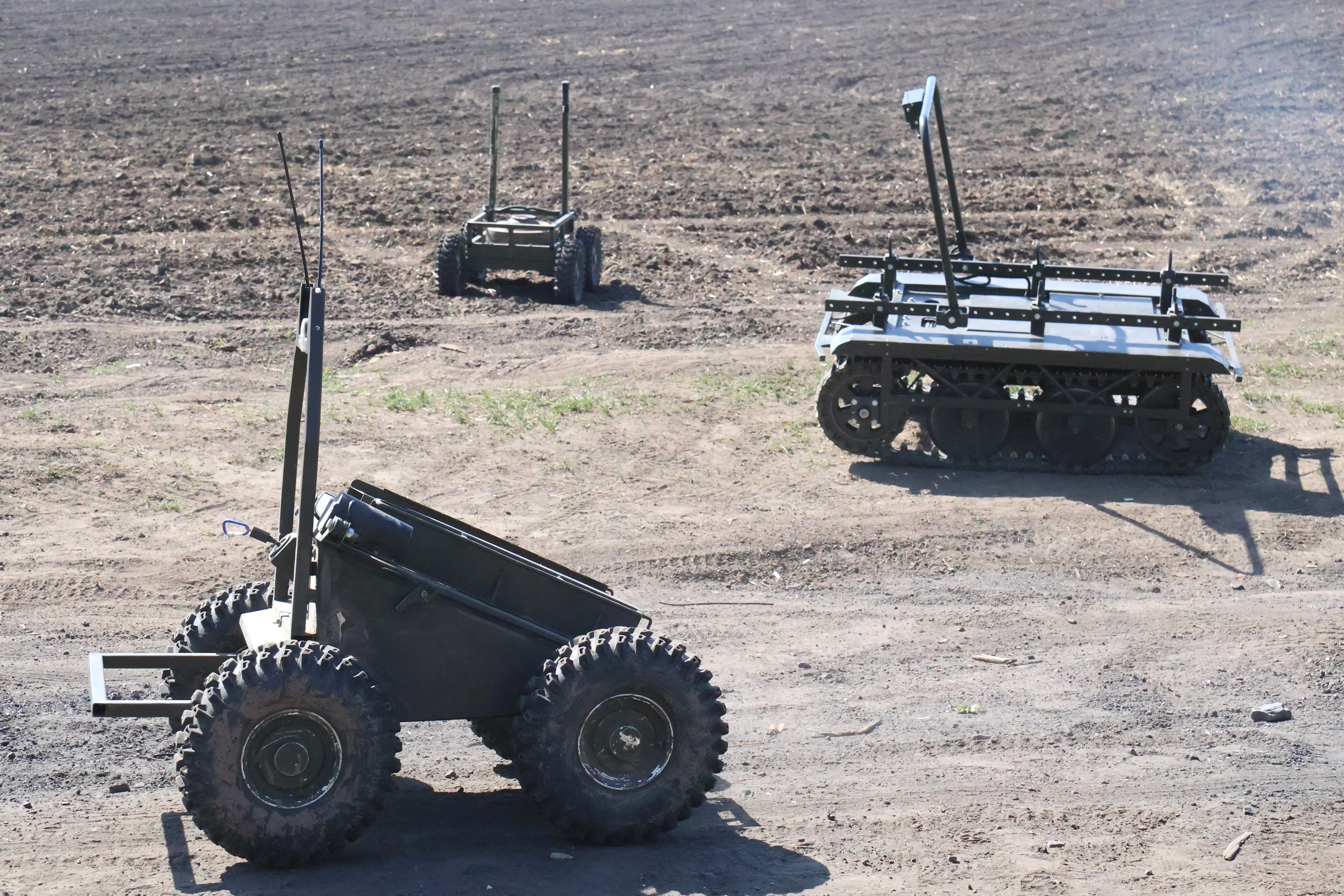 Three unmanned ground vehicles with antennas move across a dry dirt field during an outdoor demonstration.