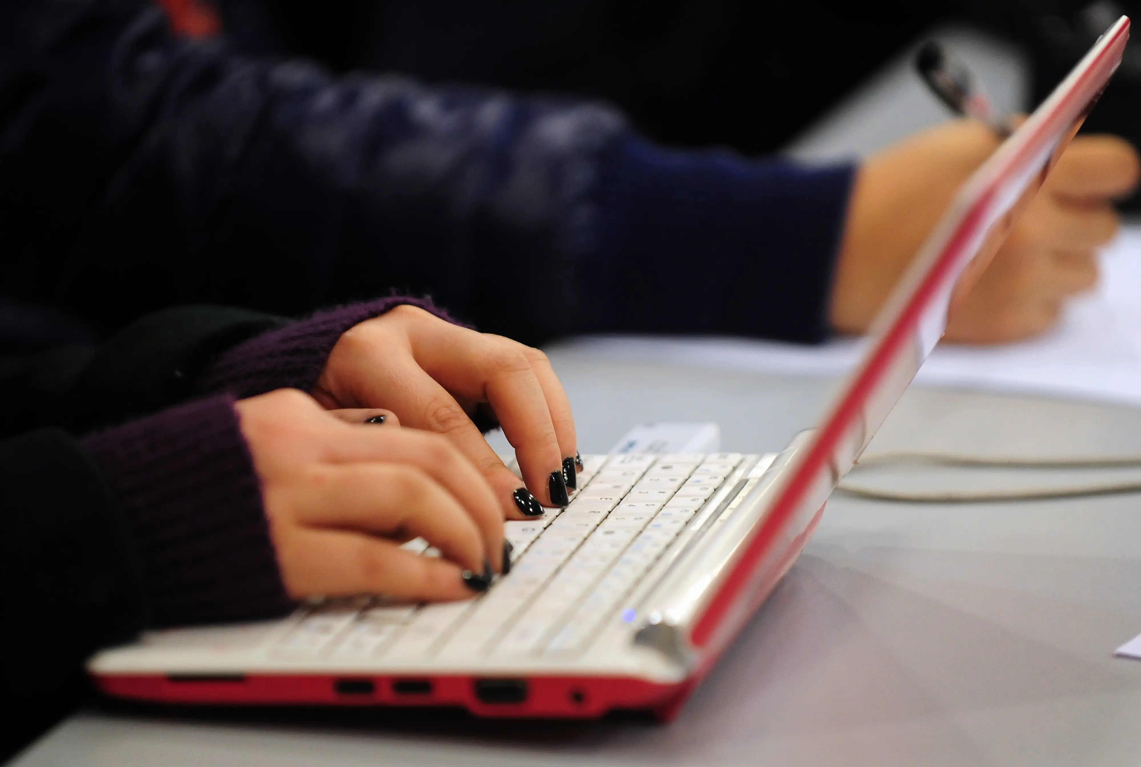 Hands type on a small pink laptop while another person writes at a shared workspace.