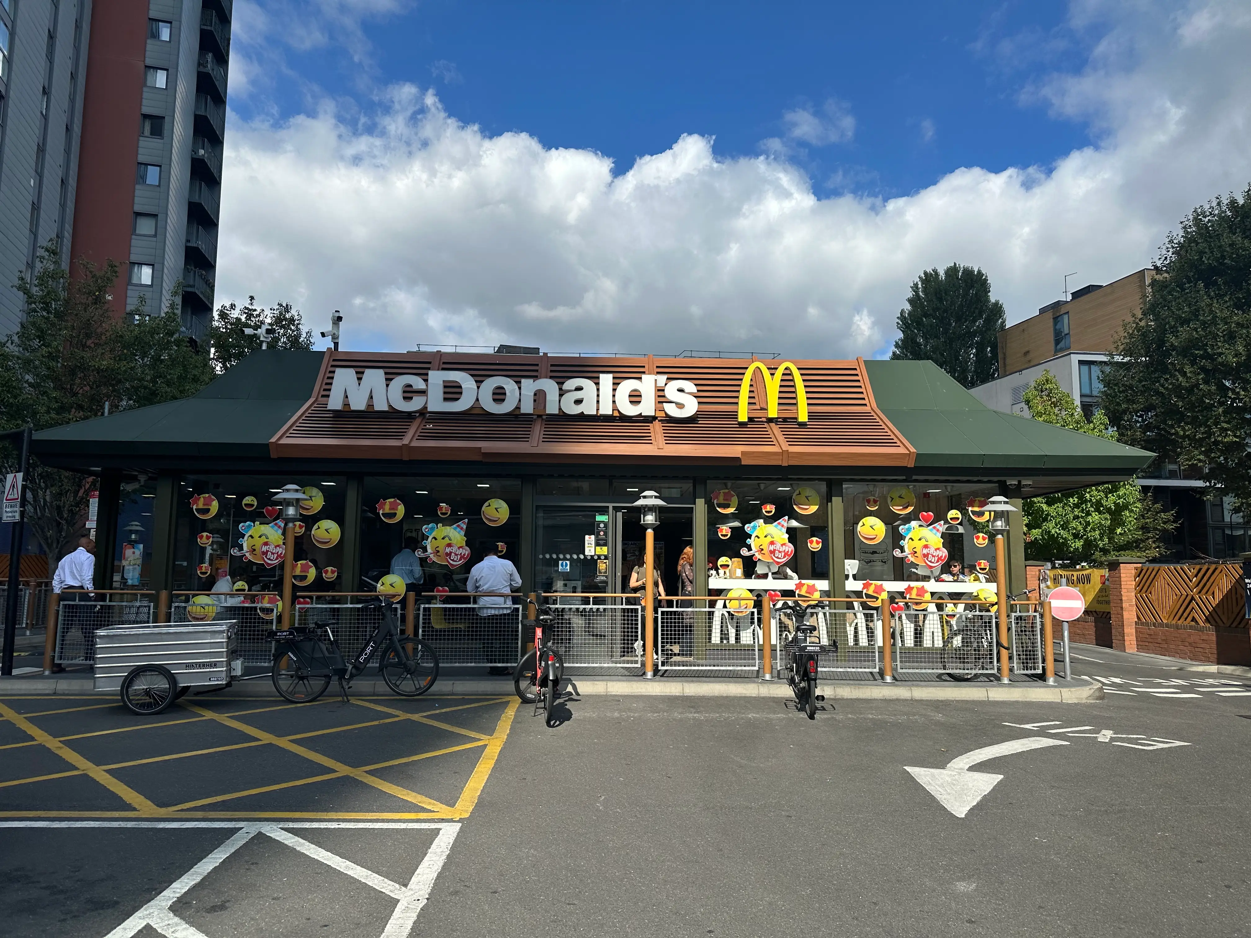 McDonald's restaurant frontage with outdoor seating, bicycles, and road markings under a partly cloudy sky.