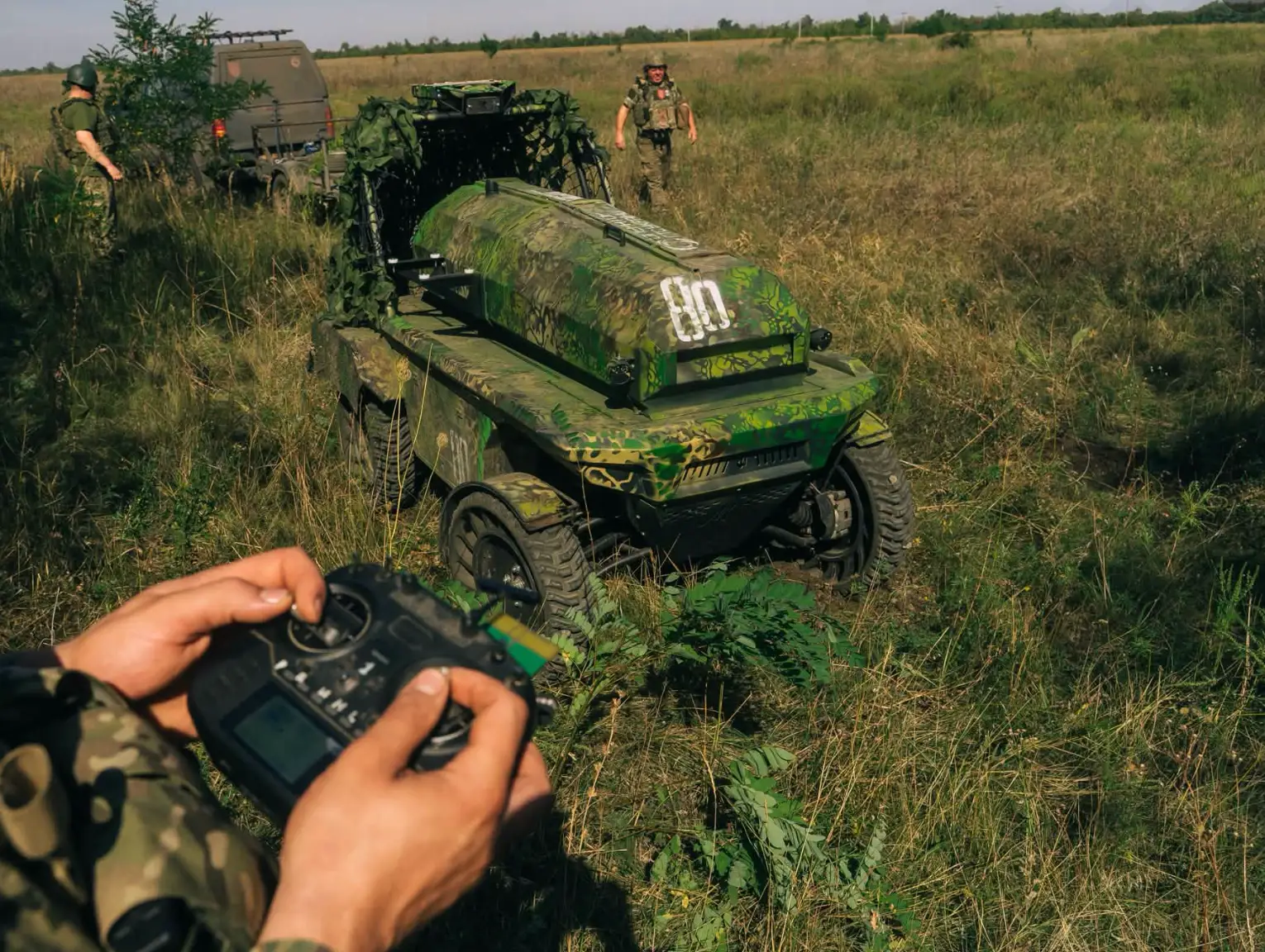 Camouflaged military personnel operate a tracked unmanned ground vehicle in a grassy field.