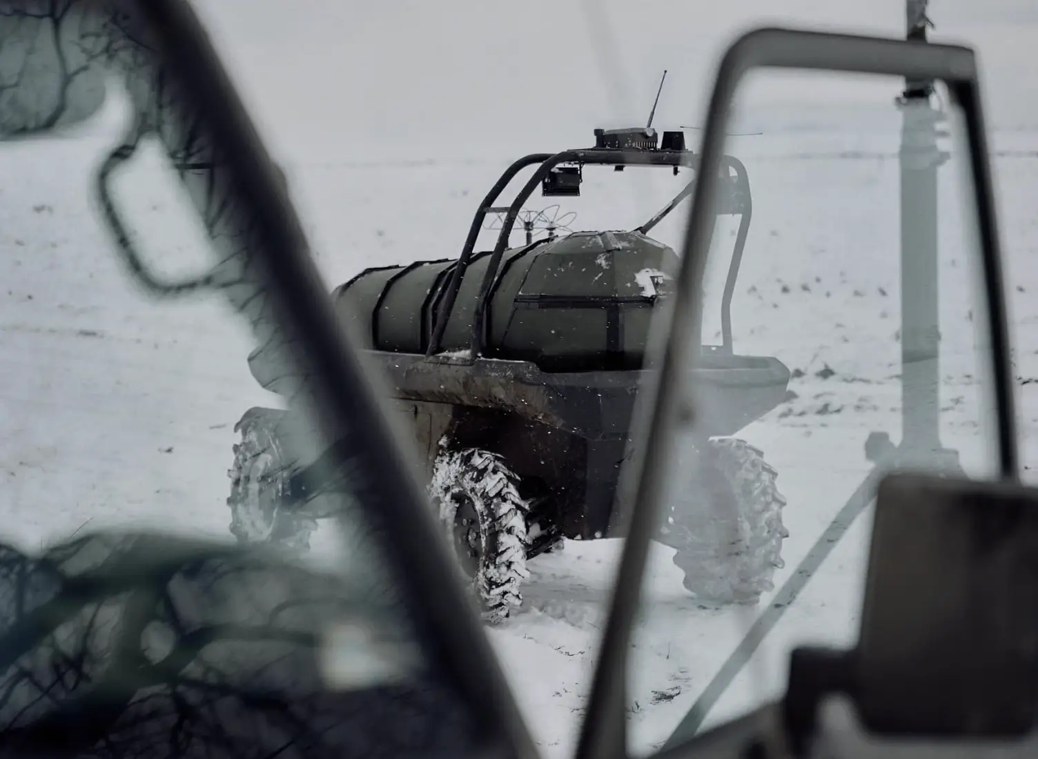 Small rugged unmanned vehicle with antennas drives across snow, viewed through a nearby vehicle window.