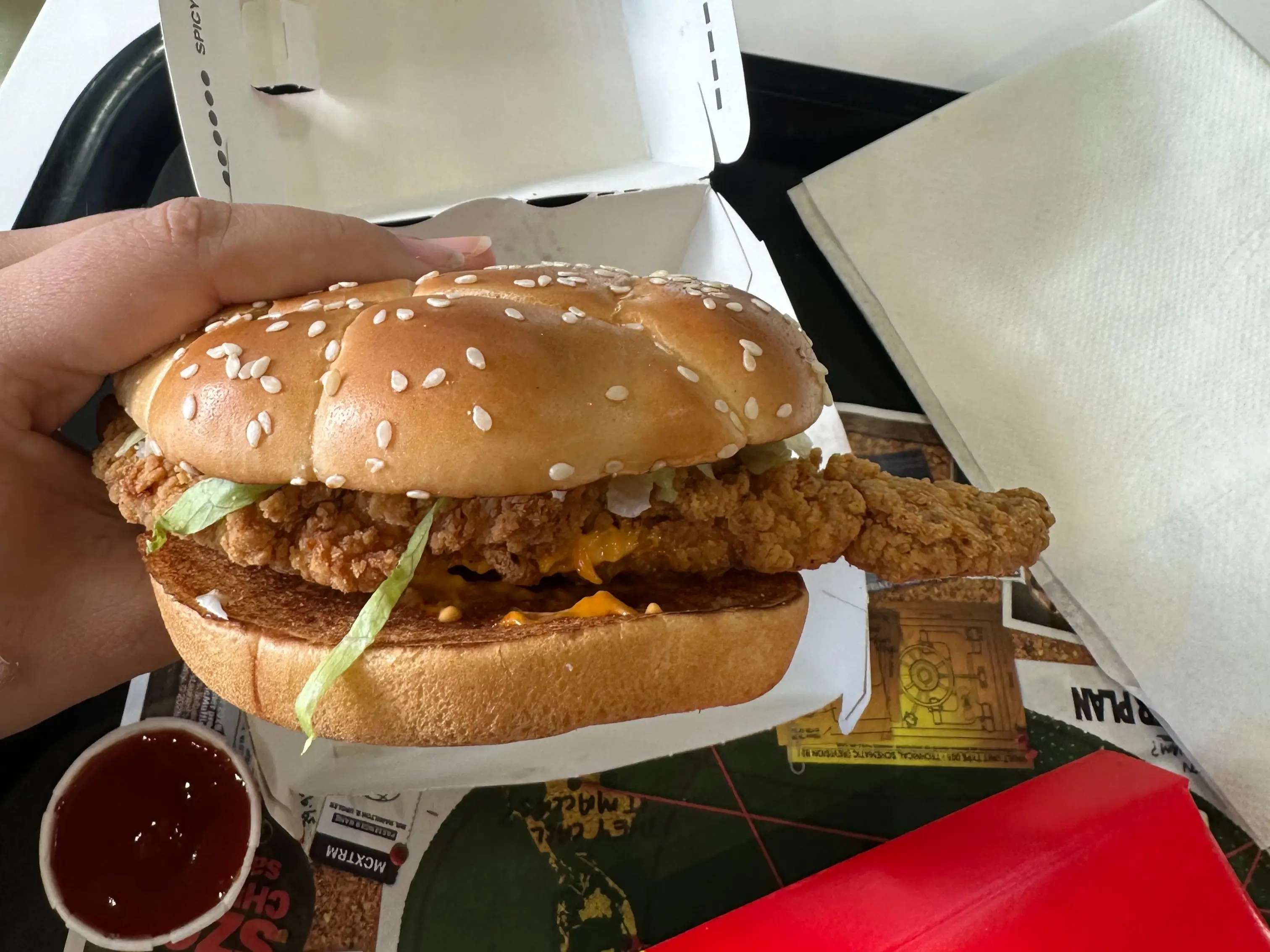 Hand holds a fried chicken sandwich with sesame bun over a fast-food tray with ketchup and napkins.