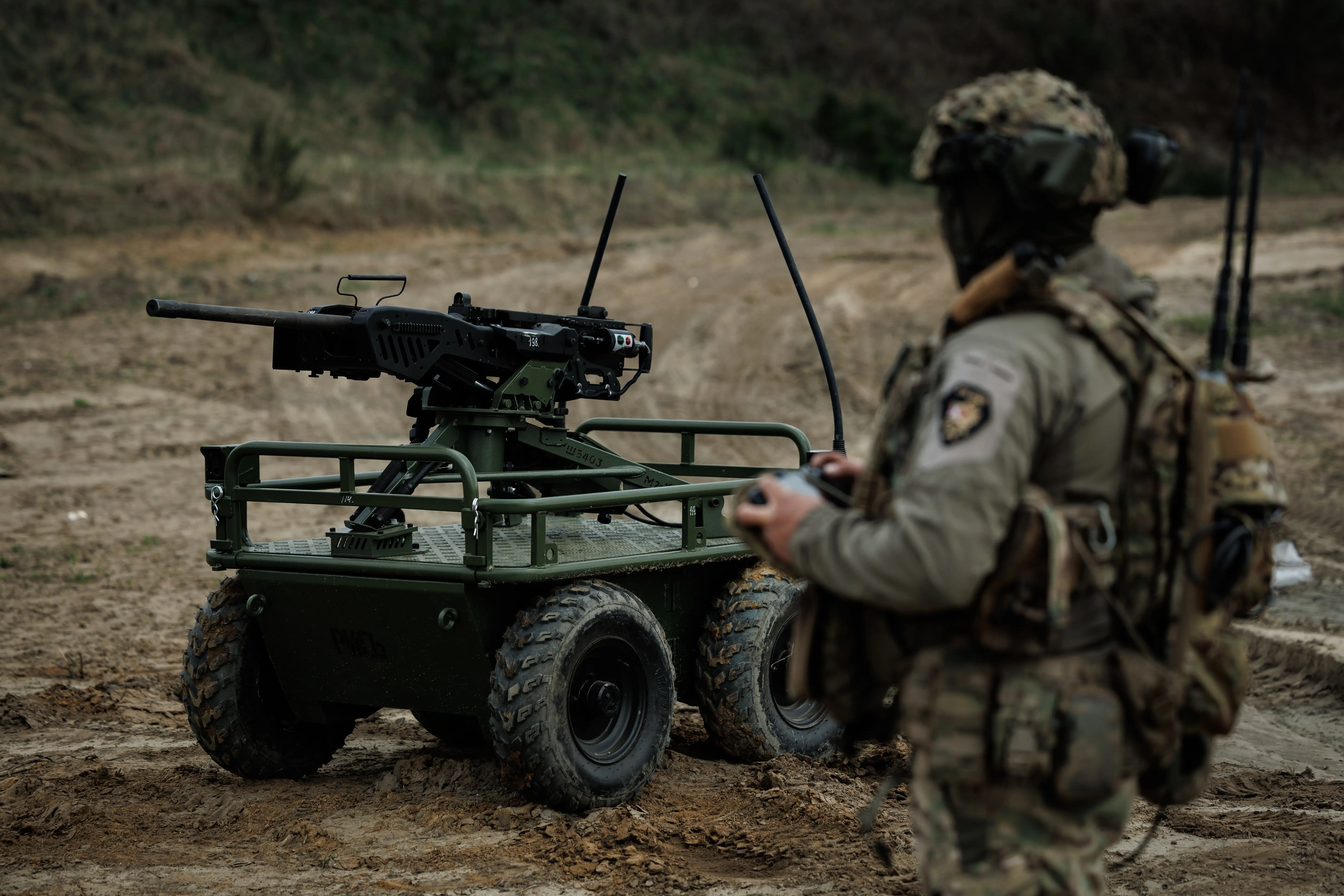 Camouflaged service member operates a weapon-mounted unmanned ground vehicle on a muddy outdoor training range.