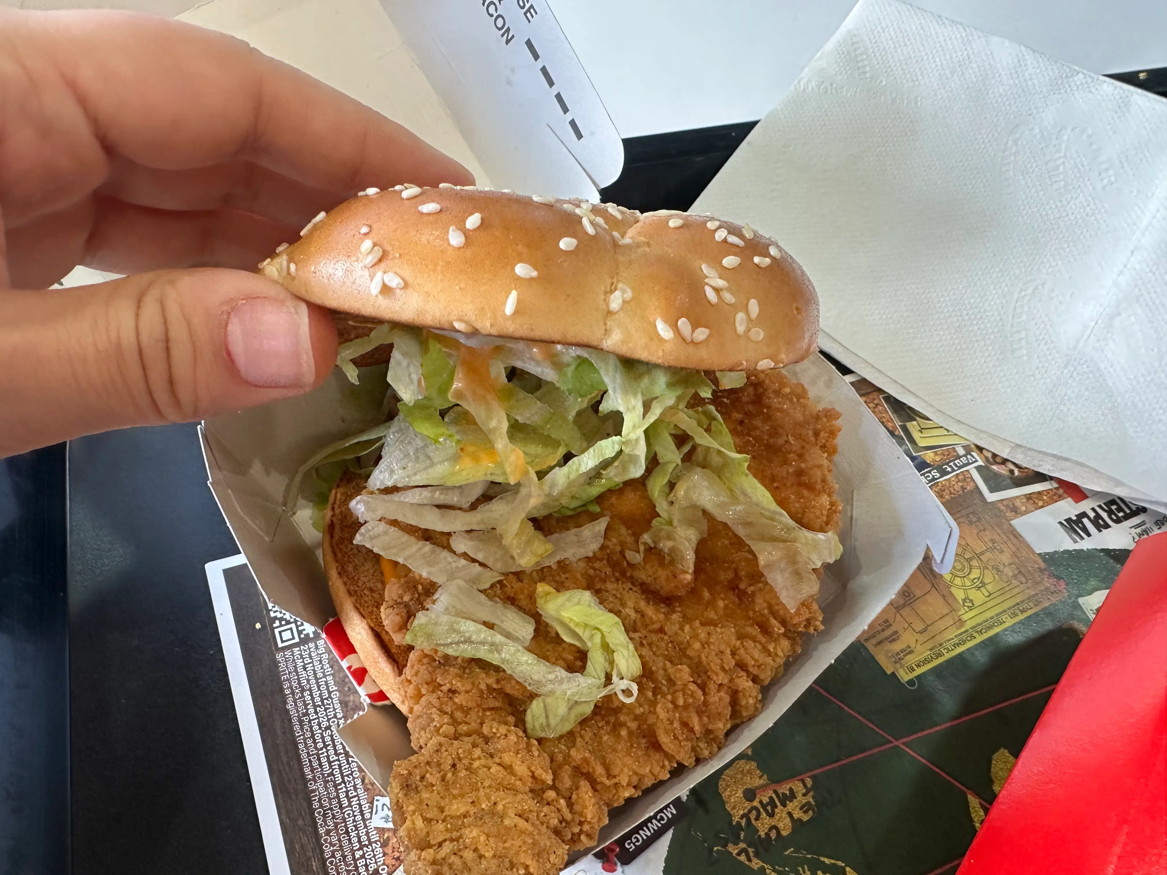A hand lifts a sesame bun on a fried chicken sandwich with lettuce in a fast food box.