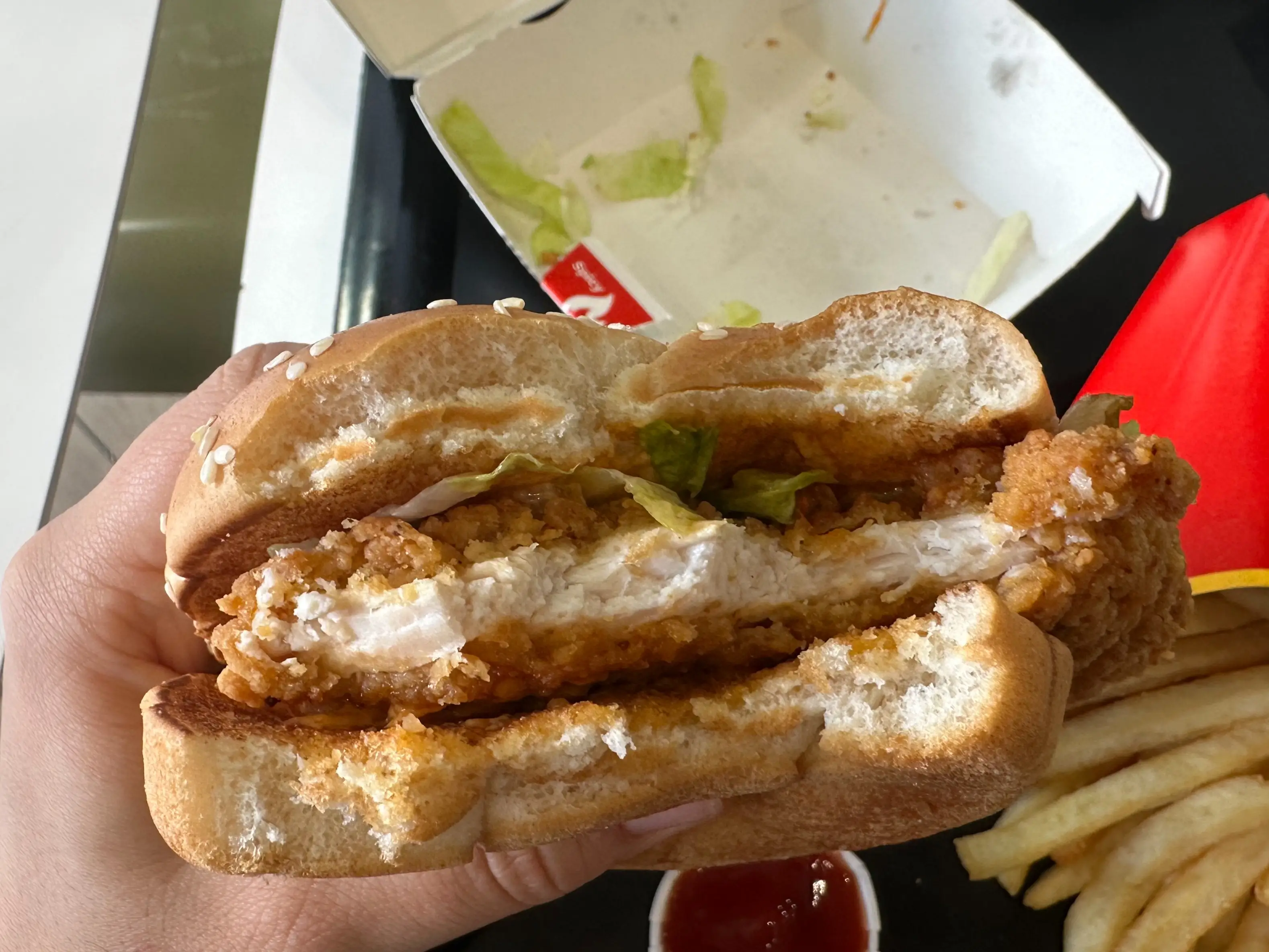 Bitten fried chicken sandwich held over a fast-food tray with fries, ketchup, and an open box.