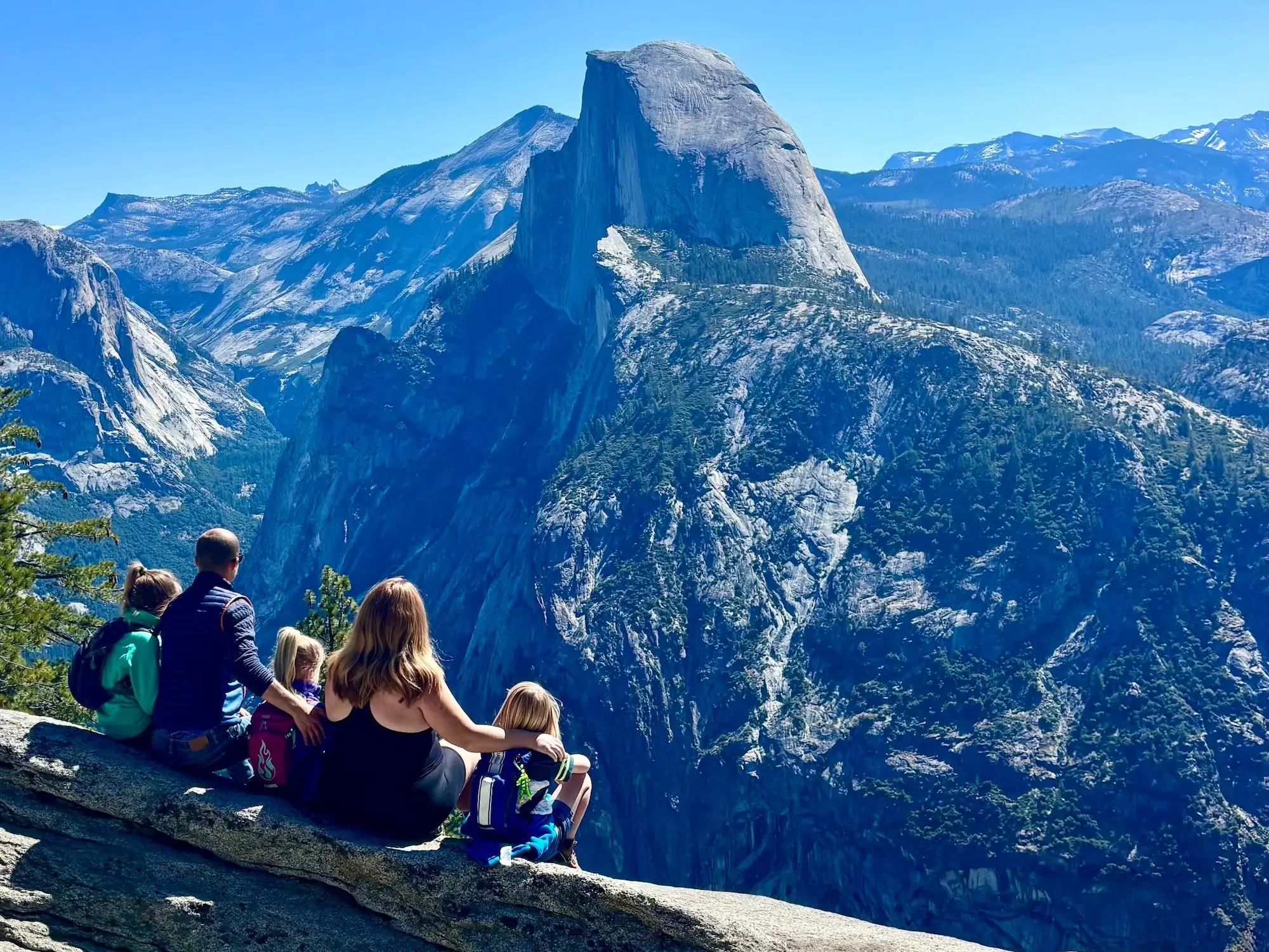 Group of visitors sits on a granite overlook facing Half Dome in Yosemite under a clear blue sky.