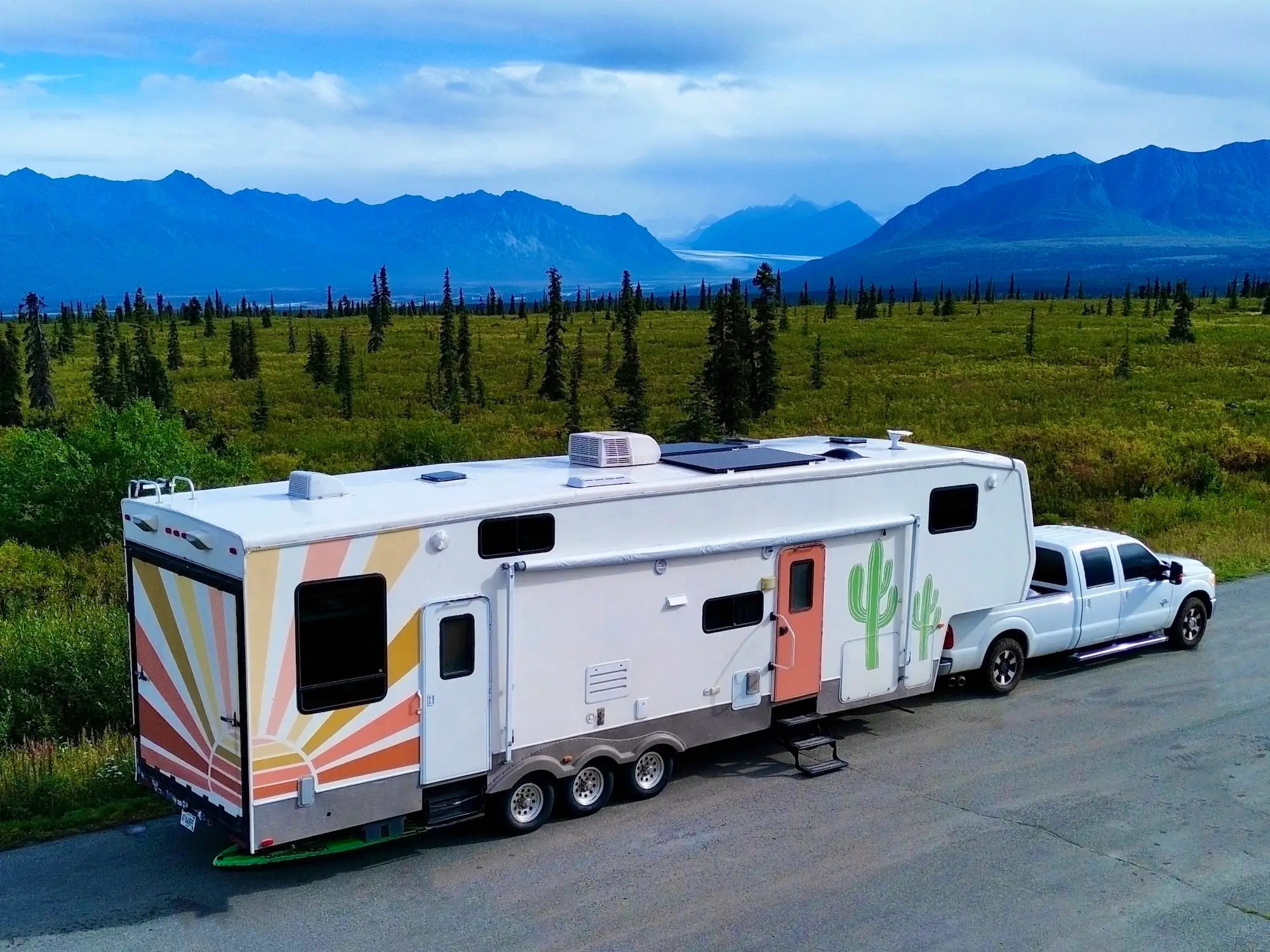 White pickup truck towing a large travel trailer on a road through green wilderness with distant mountains.