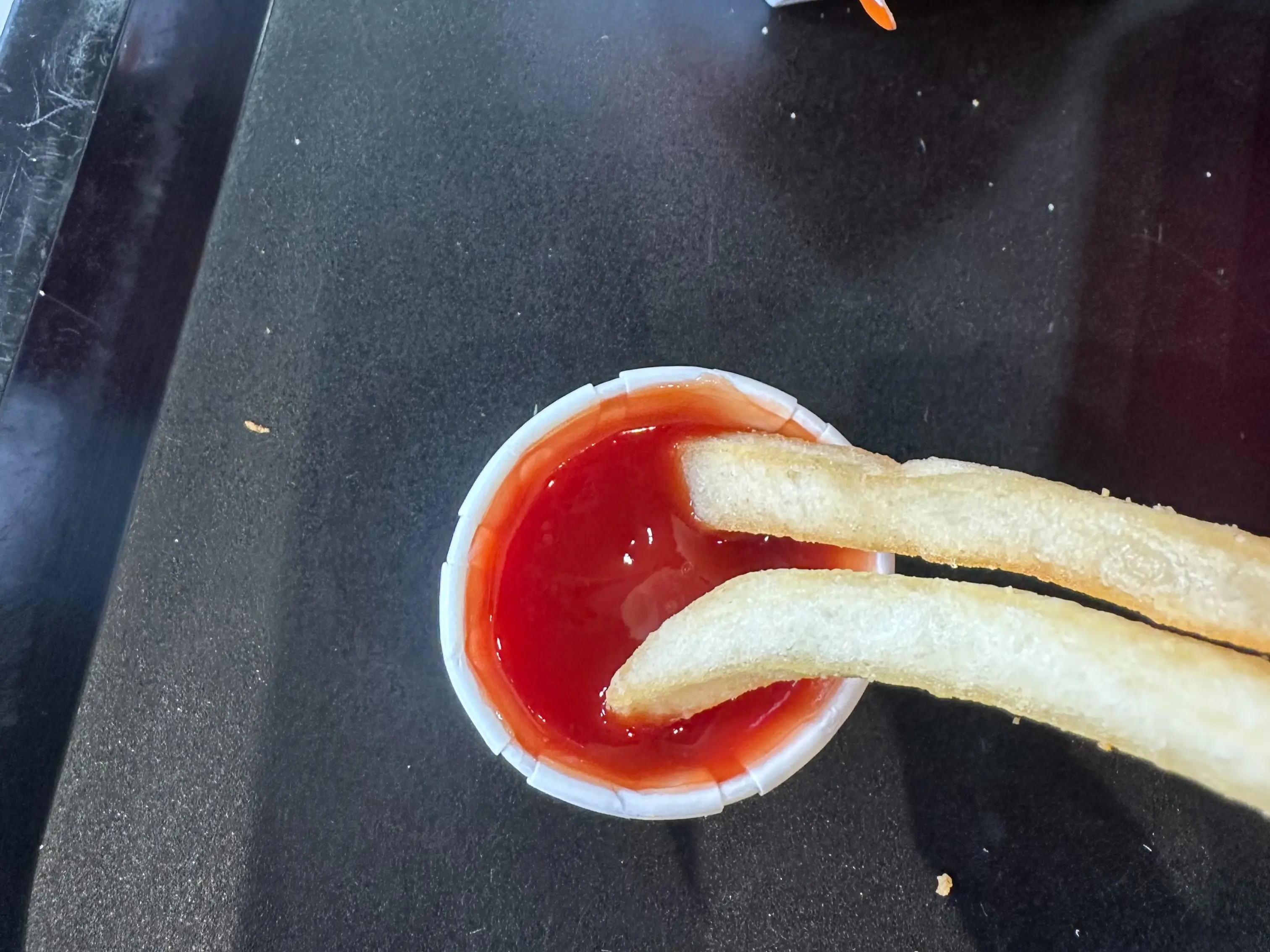 Two french fries dip into a small cup of ketchup on a black plastic tray.