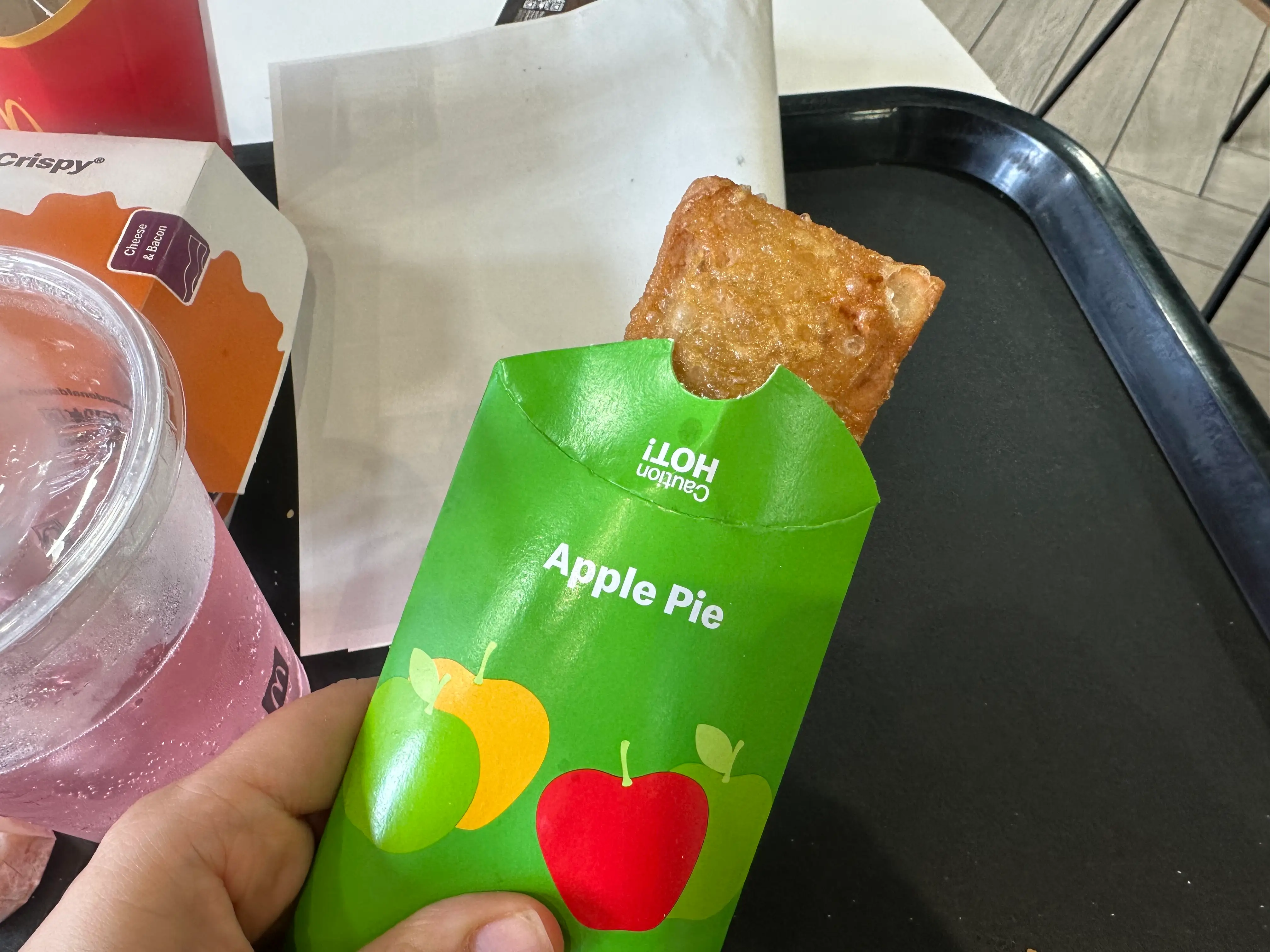 Hand holds a McDonald's Apple Pie in green packaging over a black fast food tray with a drink nearby.
