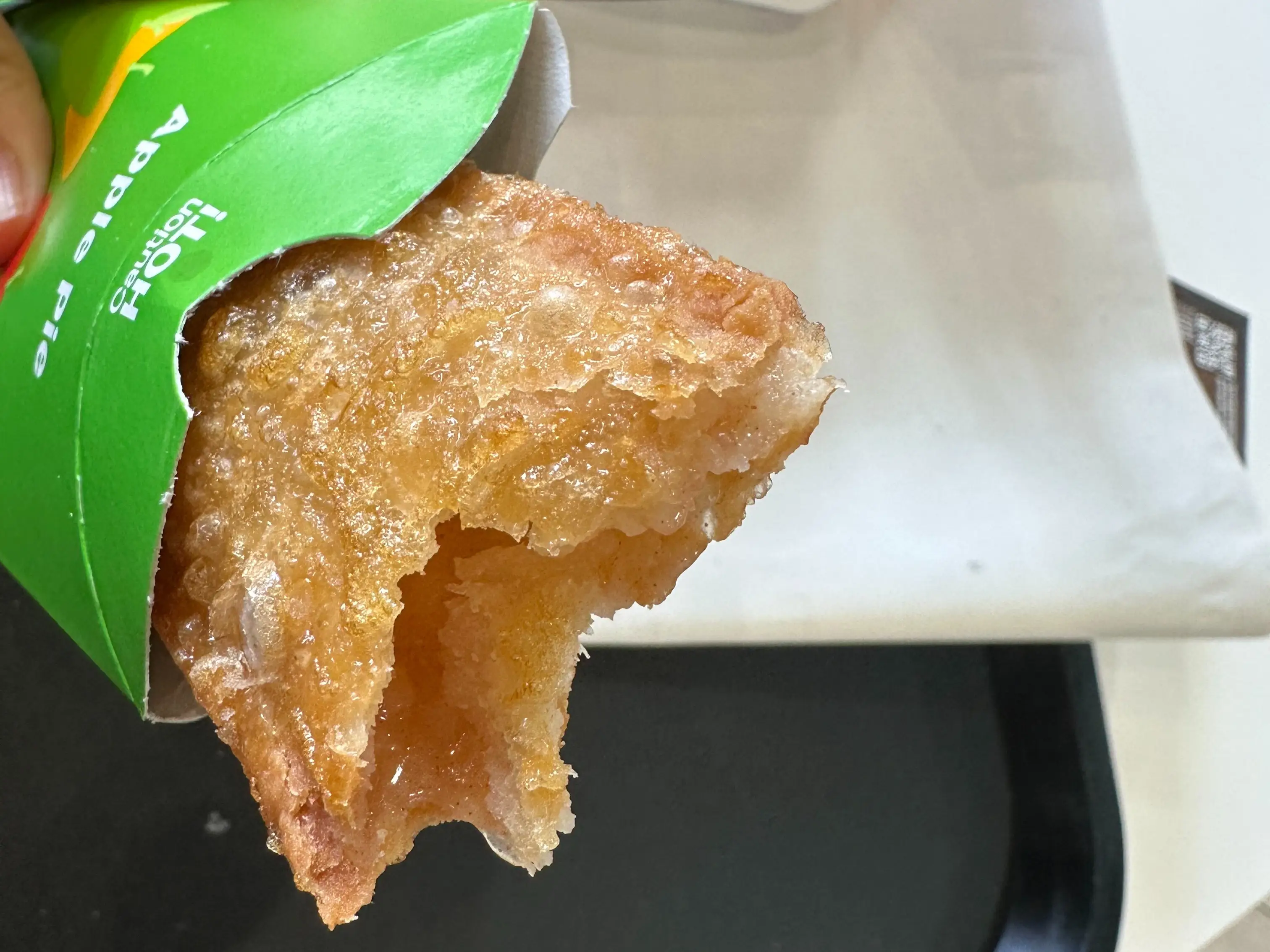 Bitten apple pie protrudes from a green Apple Pie sleeve beside a white paper bag on a tray.