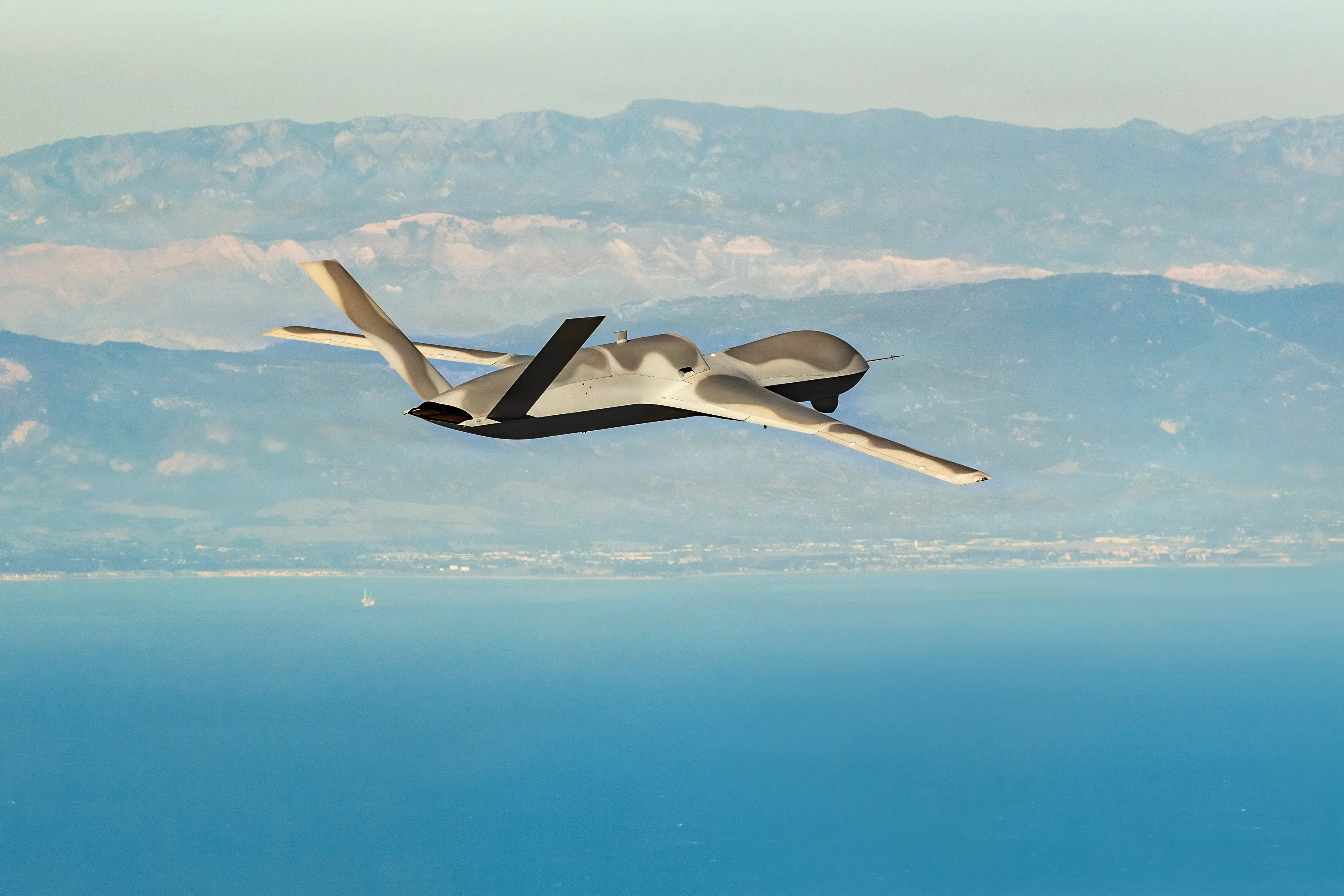 An MQ-20 Avenger uncrewed aircraft flies above a bright blue ocean with a coastal mountain range seen in the distance.